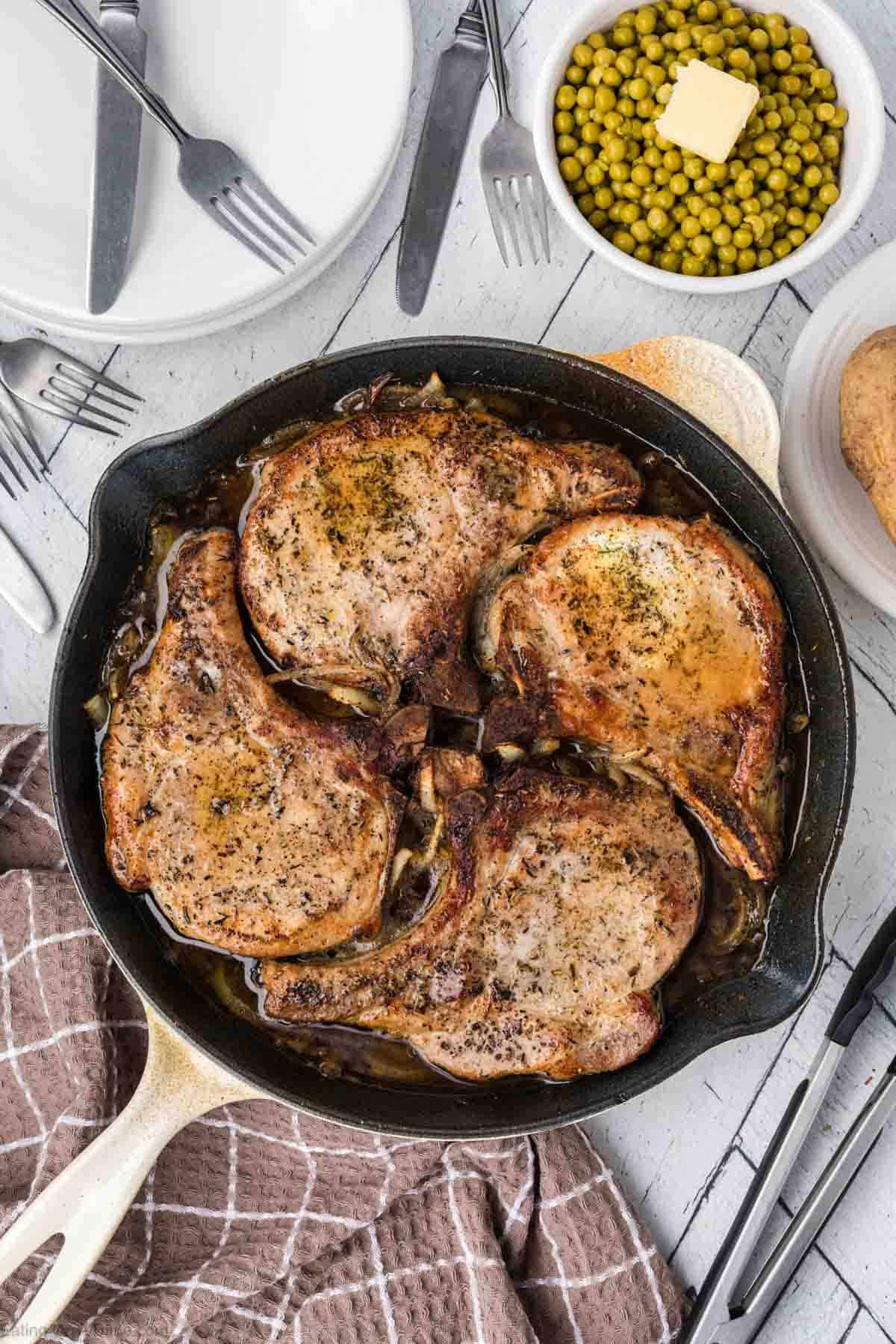 Cooked pork chops in a skillet with a bowl of peas on the side