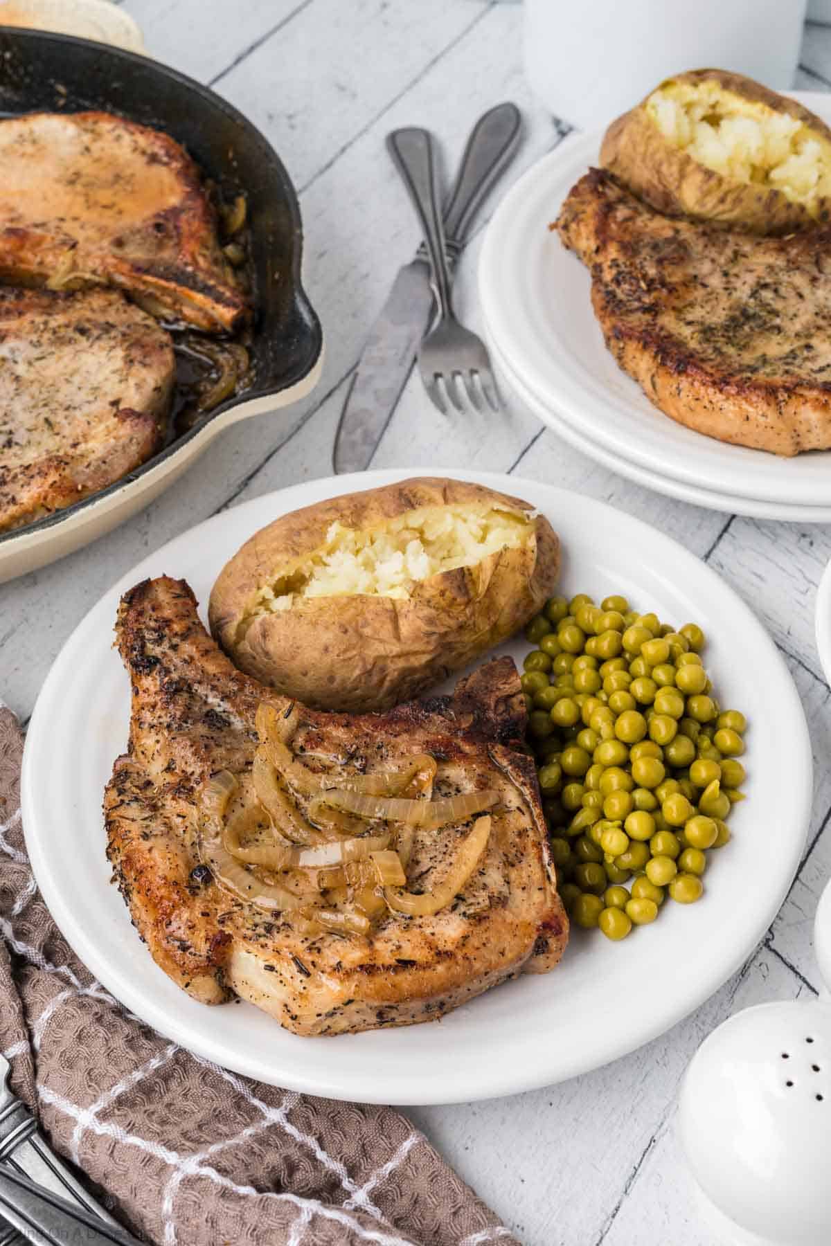 Cooked Pork Chops with caramelized onions on a plate with a bake potato and peas