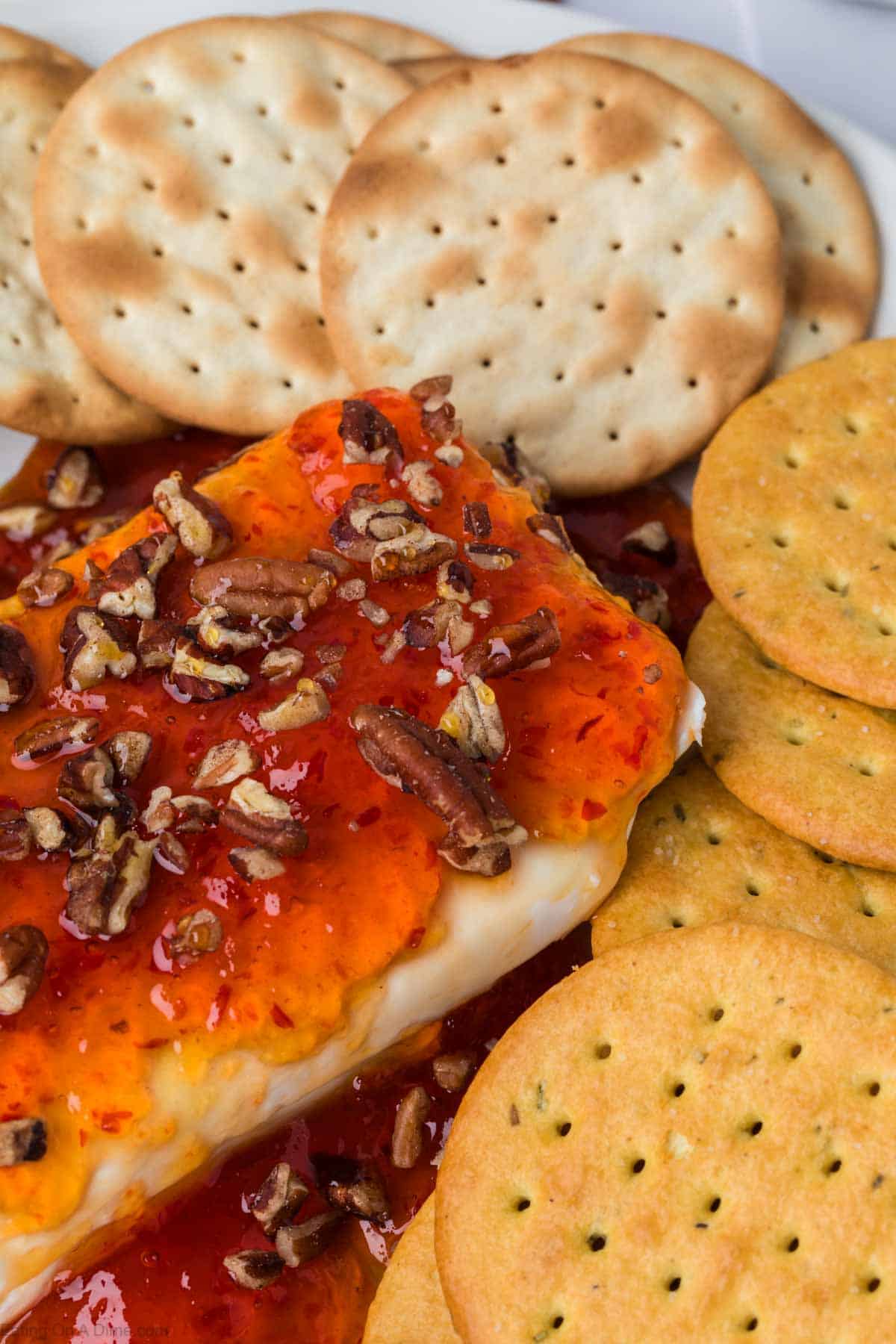 Close-up of a rich cream cheese block crowned with vibrant pepper jelly and chopped pecans, encircled by round crackers. This colorful appetizer creates a delightful contrast against the light and golden hues of the crispy crackers.