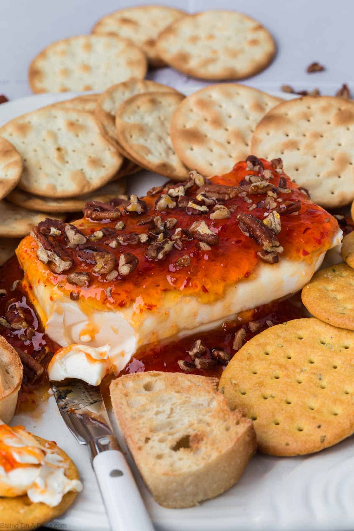 A block of cream cheese topped with zesty pepper jelly and chopped pecans is surrounded by a variety of crackers and small bread slices on a white plate. A spreader knife rests in the cheese, coated with jelly and nuts, ready for spreading.