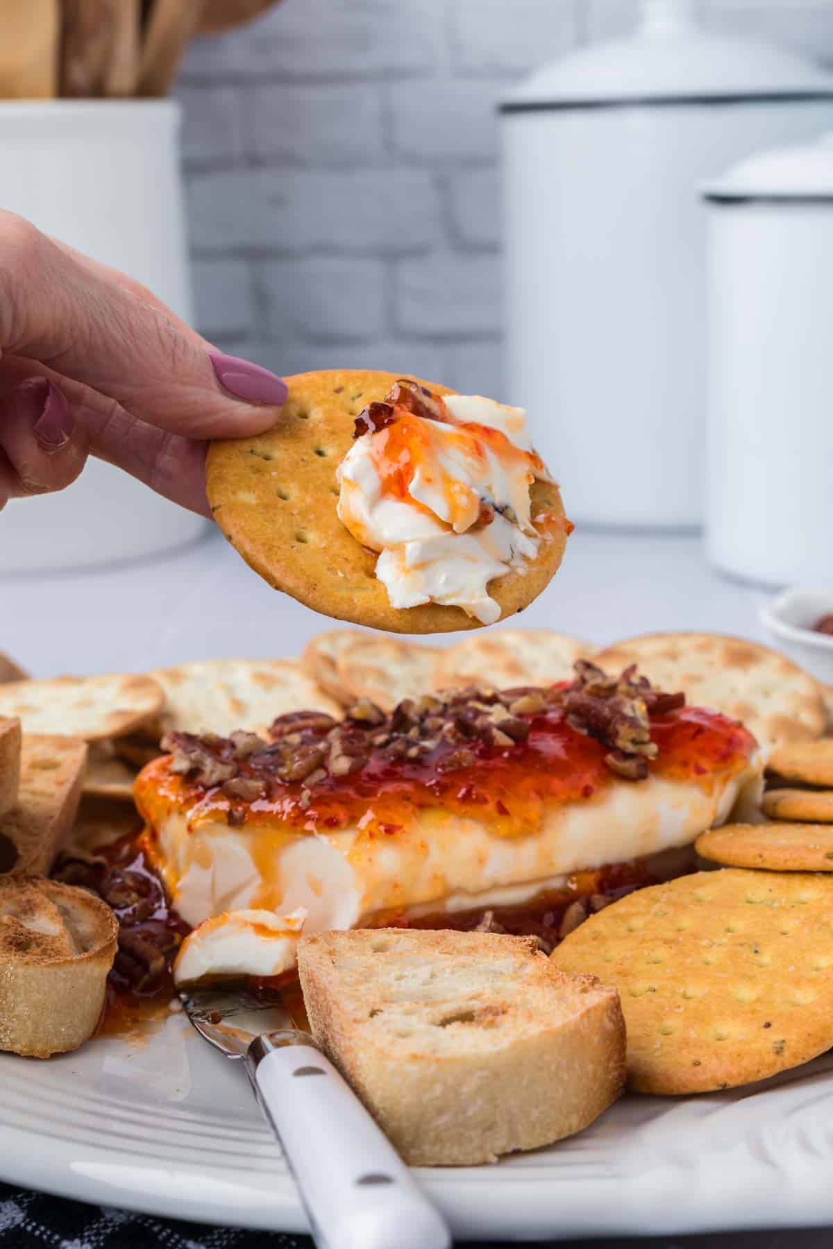 A person holds a cracker topped with creamy cheese, drizzled with vibrant pepper jelly. The serving plate below features cream cheese, pecans, the fiery sauce, and an assortment of crackers and bread slices. A spreader lies on the plate.