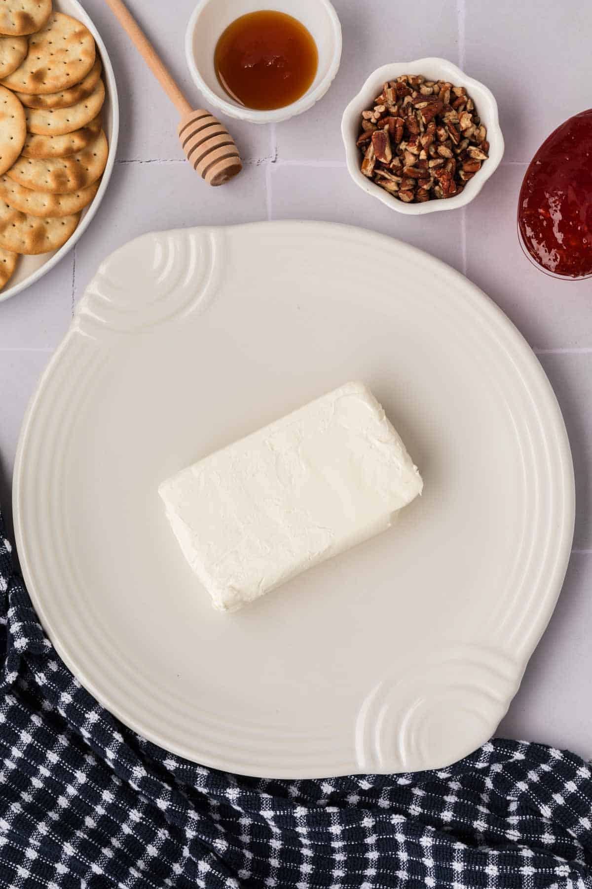 A block of cream cheese sits on a decorative plate, surrounded by a small dish of honey with a dipper, chopped pecans, and crackers. A bowl of pepper jelly adds a spicy twist, while a black and white checkered cloth is partially visible at the bottom.