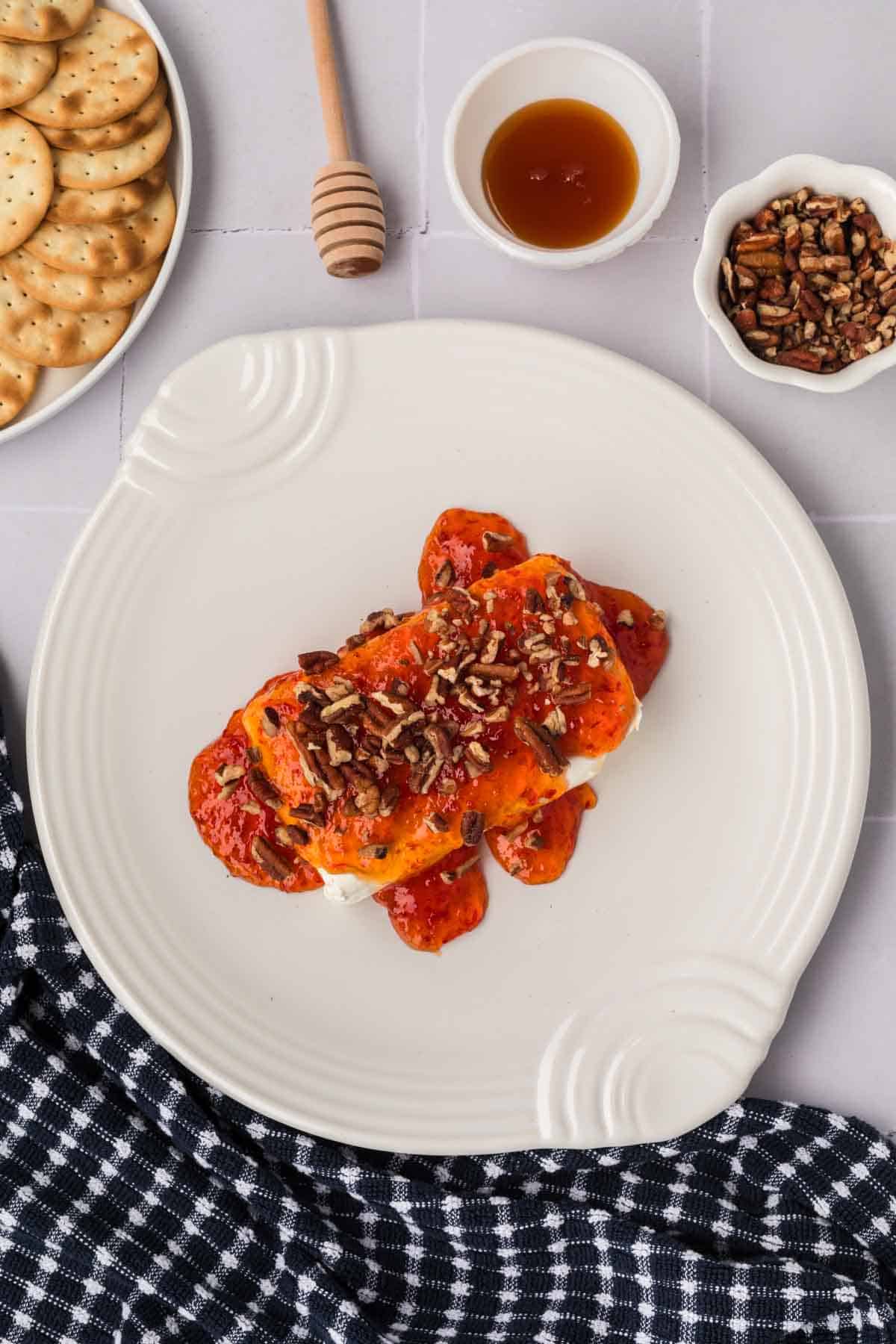 A plate with cream cheese crowned by tangy pepper jelly and chopped pecans. Nearby, a small bowl of honey and a bowl of pecans accent the spread, complemented by a plate of crackers on a checkered cloth.