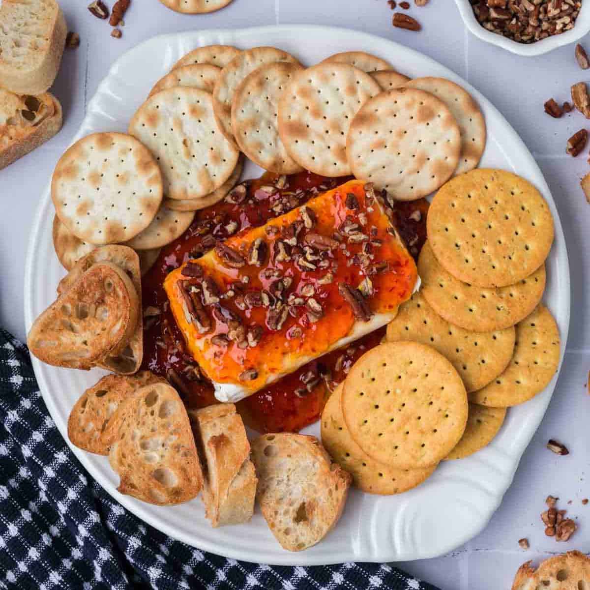 A plate topped with a rectangle of cream cheese and pepper jelly, adorned with pecan pieces. It’s surrounded by round crackers, toasted bread slices, and rests on a classic black-and-white checkered cloth, creating a delightful centerpiece for any gathering.