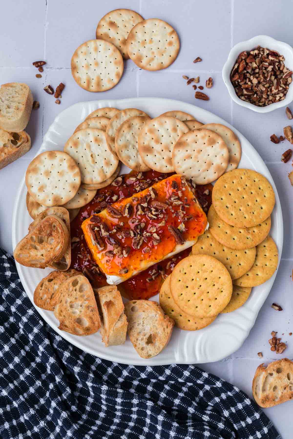 A white plate with crackers and sliced bread surrounds a block of cream cheese topped with red pepper jelly and pecans. A small bowl of pecans and a checkered cloth are nearby, completing this delightful spread.