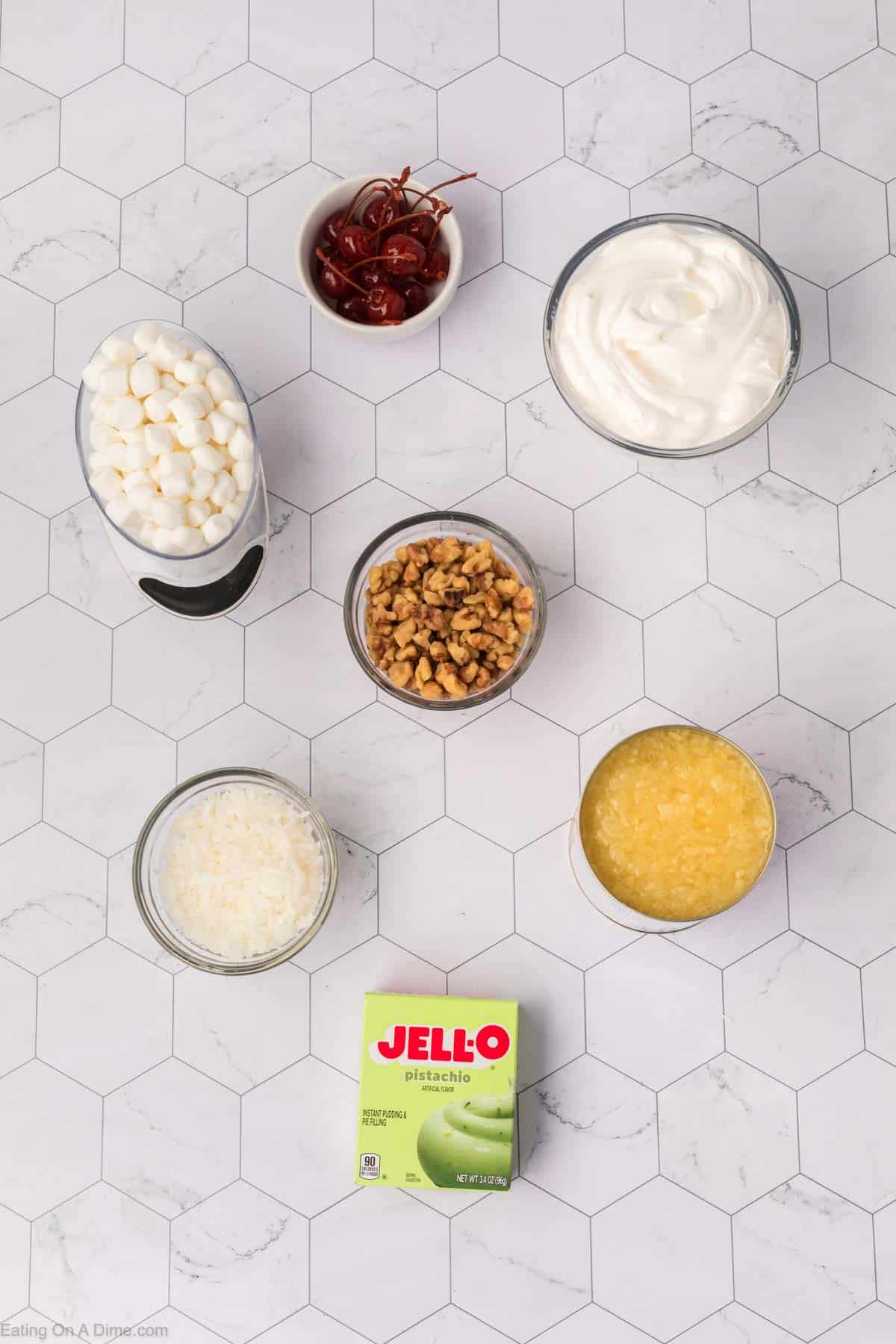 A hexagonal tile surface showcases the delightful ingredients for a classic Watergate Salad: a bowl of whipped cream, canned pineapple, chopped walnuts, shredded coconut, marshmallows, maraschino cherries, and a box of Jell-O pistachio pudding mix—a timeless dessert recipe.