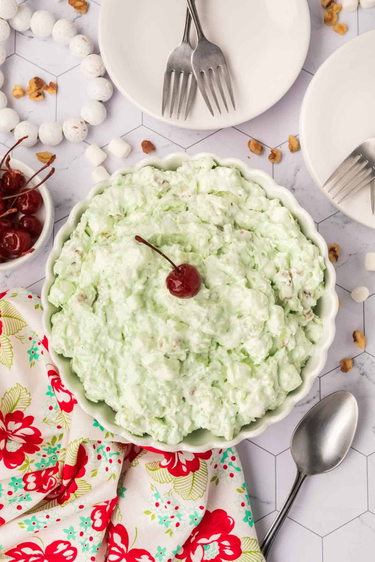 A bowl of luscious Watergate Salad, adorned with a cherry, sits amid forks, a vibrant floral napkin, and scattered nuts and marshmallows atop a hexagonal-patterned surface.