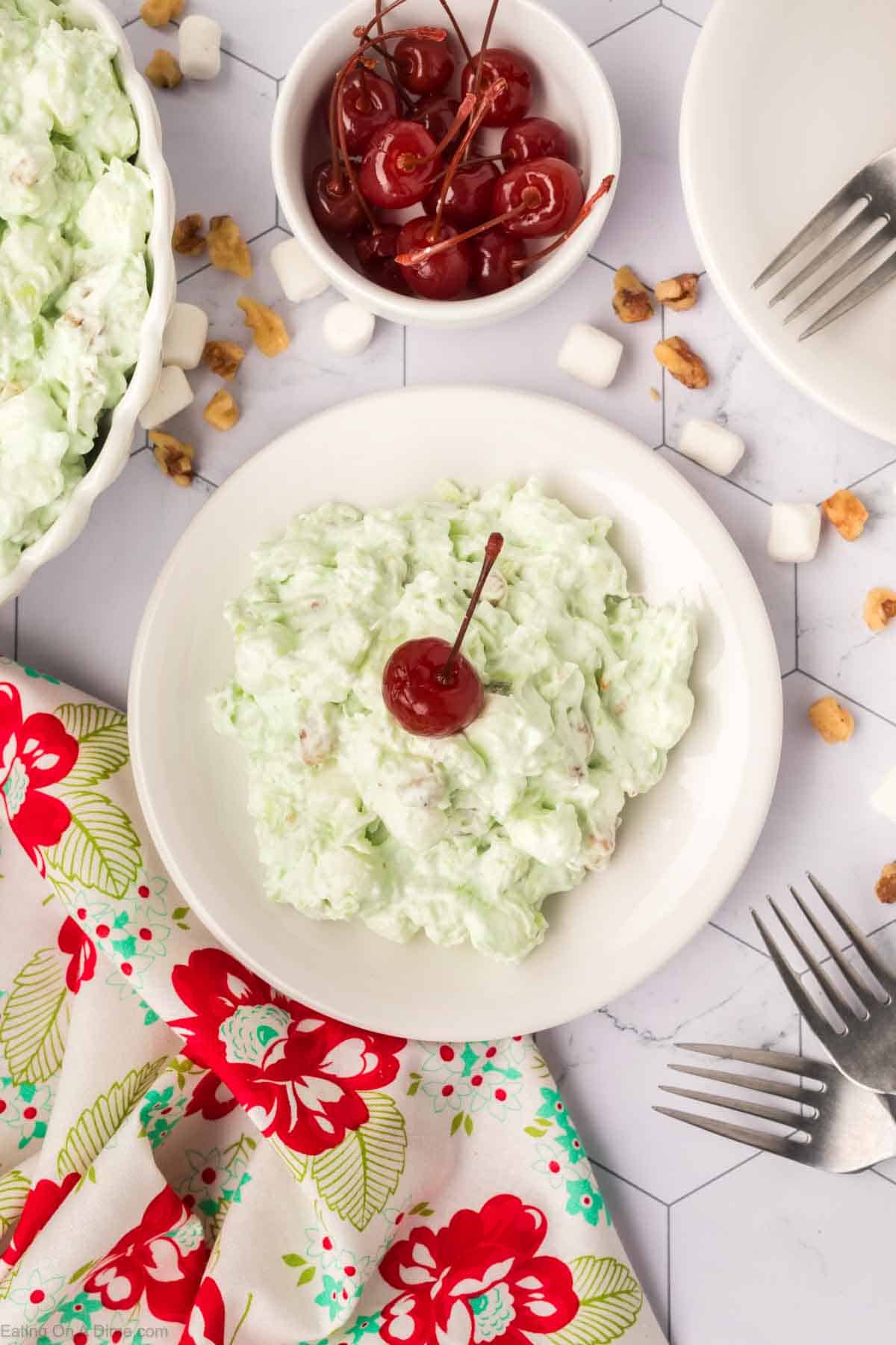 A white plate holds a serving of the classic Watergate Salad, a delightful dessert featuring green pistachio flavors topped with a cherry. Nearby, a bowl of cherries and mini marshmallows rest on a colorful floral napkin, while two forks lie poised on the patterned tablecloth.