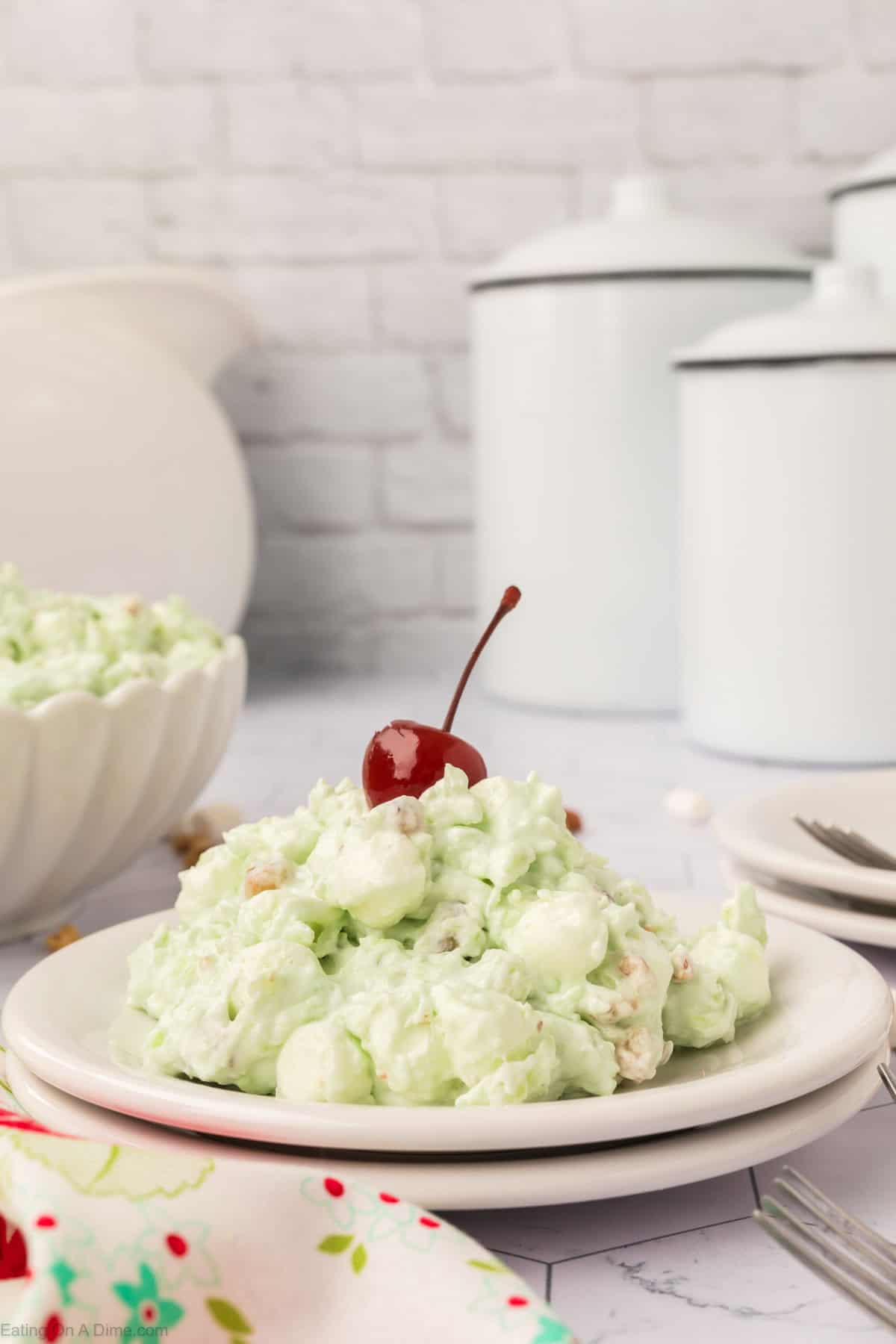 A plate of vintage Watergate Salad garnished with a cherry on top. This classic salad is fluffy, featuring marshmallows and nuts. In the background, white canisters and a bowl with more salad rest on a pristine white countertop, evoking timeless charm from traditional salad recipes.