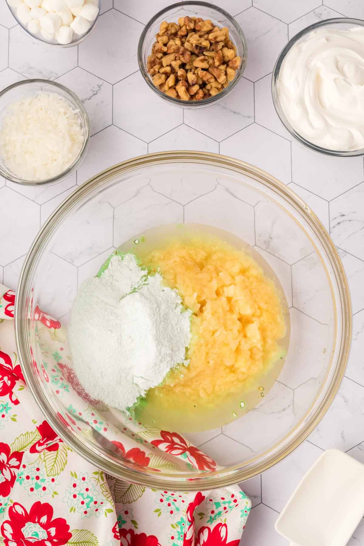 A large glass bowl with lime green gelatin mix and crushed pineapple sits on a hexagonal patterned surface, reminiscent of a classic Watergate Salad. Surrounding it are small bowls of marshmallows, nuts, shredded coconut, and whipped cream. A floral cloth is partially visible.