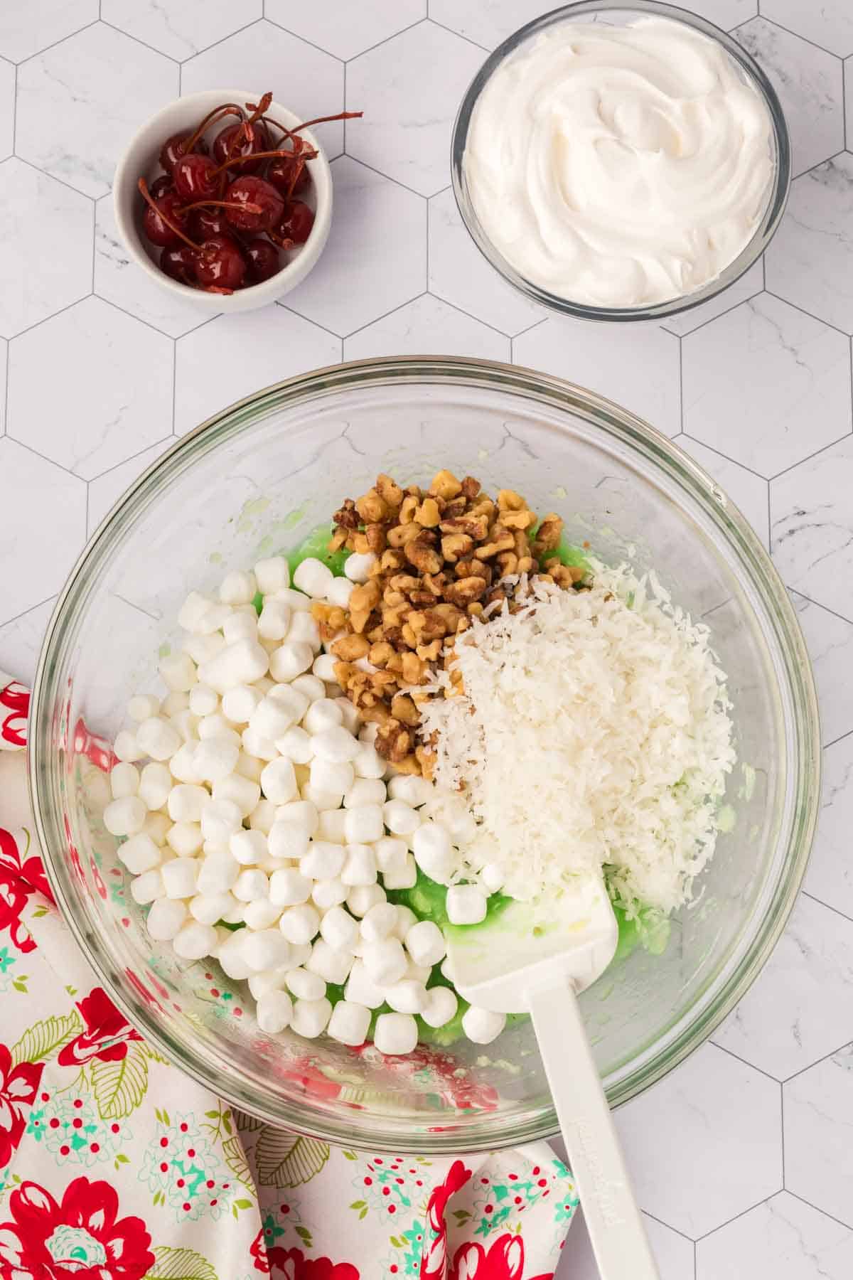 A glass bowl on a hexagonal white surface holds the classic Watergate Salad ingredients: mini marshmallows, chopped walnuts, and shredded coconut atop a green pistachio mixture. Nearby bowls overflow with whipped cream and maraschino cherries, while a floral cloth adds a touch of elegance.