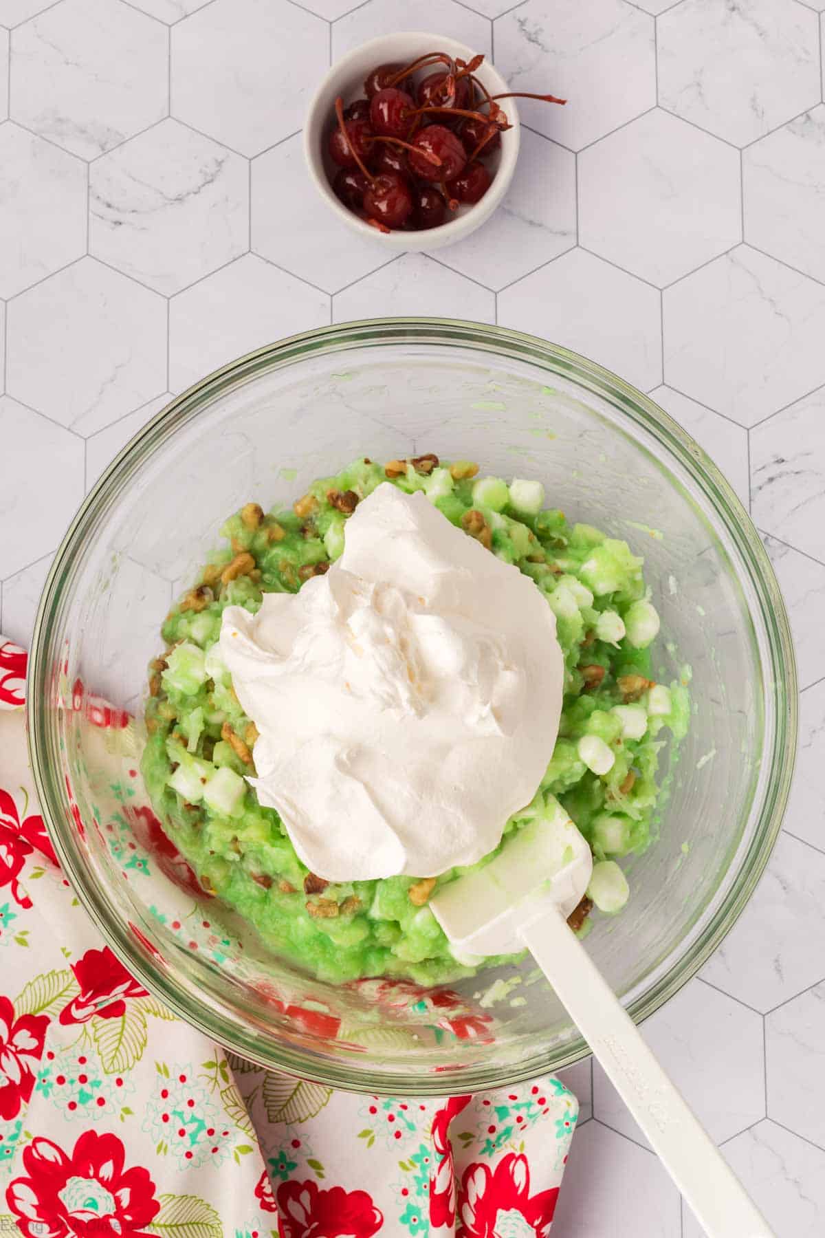 A bowl of pistachio pudding mix with crushed pineapple, mini marshmallows, and chopped nuts forms the delightful Watergate Salad dessert. It's topped with whipped cream, a spatula rests in the bowl. In the background, a small bowl of maraschino cherries and a floral cloth are visible.