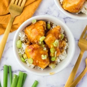 A bowl of rice topped with glazed chicken pieces, garnished with chopped green onions, graces the marble surface—a delightful addition to your Meal Plan 28. Nearby, golden forks elegantly rest on a napkin.