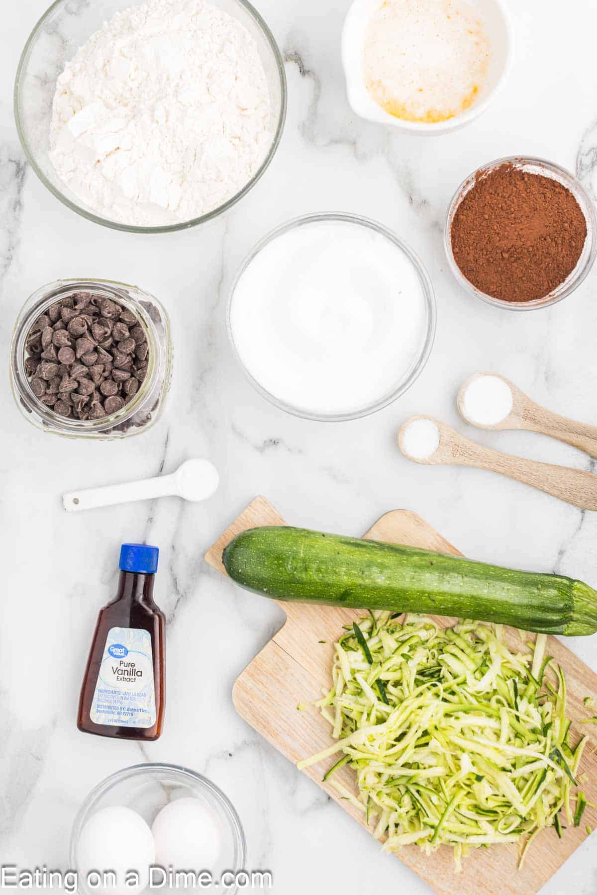 Ingredients for baking a cake are laid out on a marble surface: flour, cocoa powder, sugar, chocolate chips, and vanilla extract, resembling the beginnings of a delectable chocolate zucchini bread. A fresh zucchini is shredded on a cutting board next to the eggs and measuring spoons.