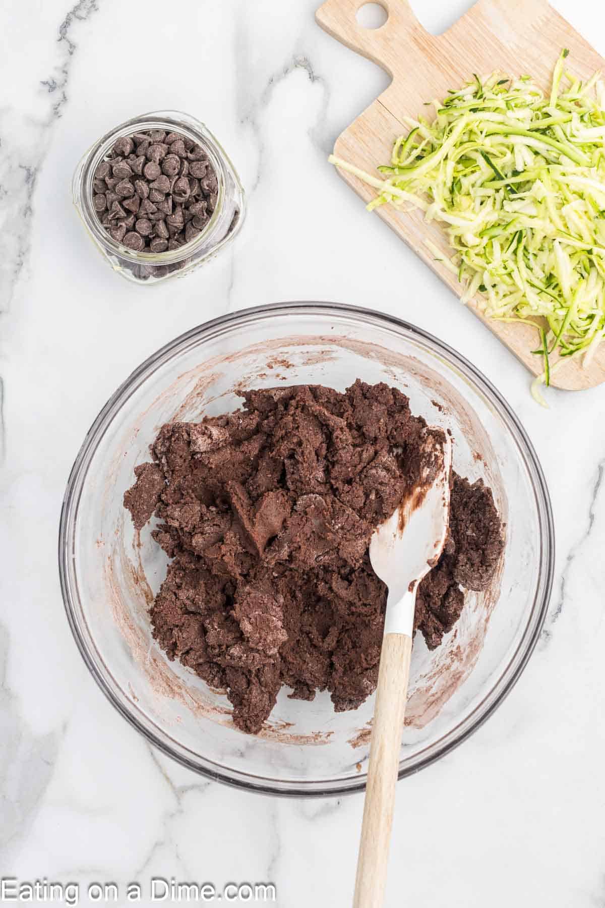 A glass bowl filled with rich chocolate cookie dough sits alongside a spatula, while a wooden board holds shredded zucchini, hinting at a delightful twist on classic bread. A small jar of chocolate chips adds temptation on the marble countertop.