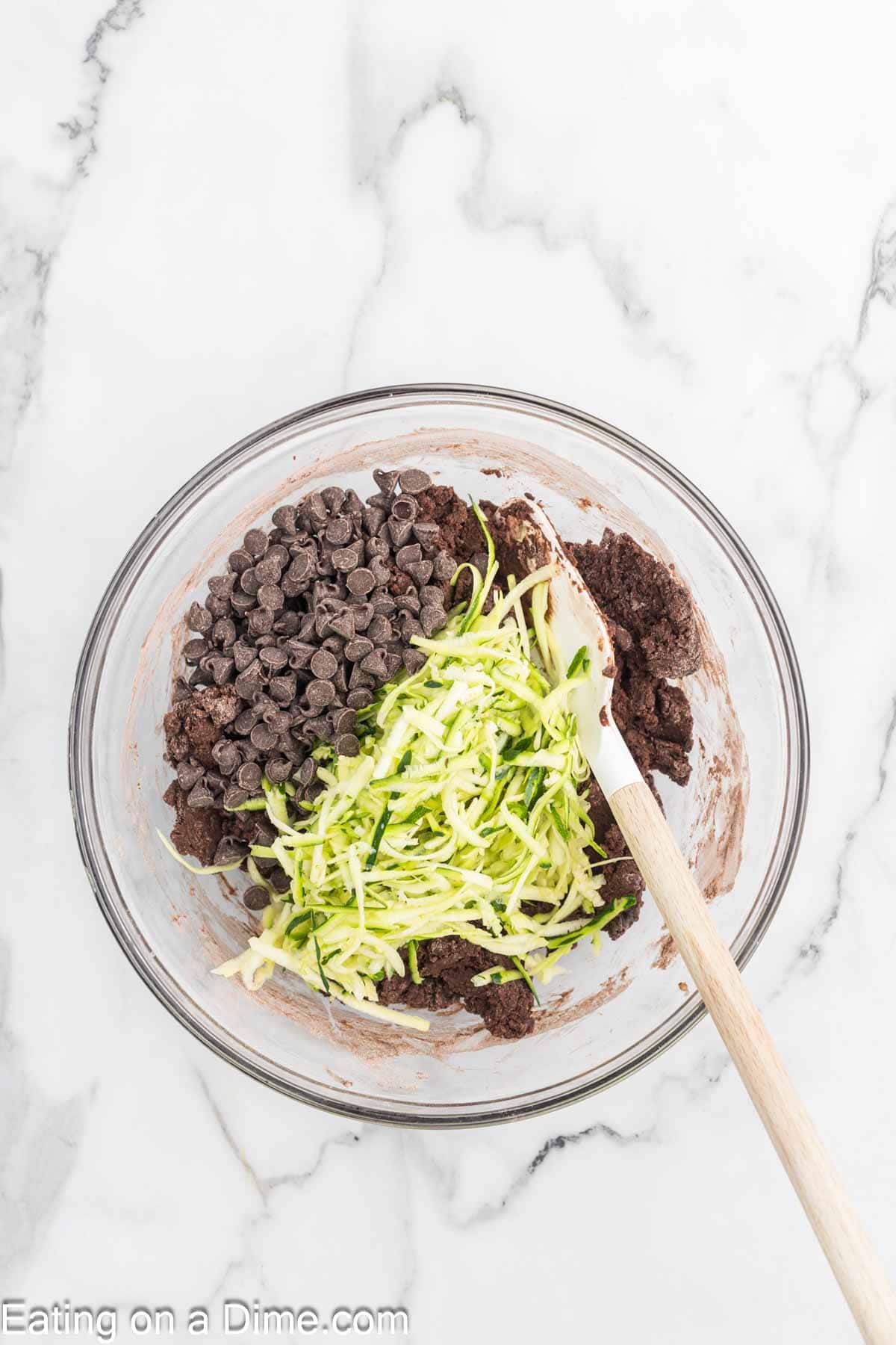 A glass bowl on a marble countertop holds rich brownie batter, chocolate chips, and shredded zucchini, poised to transform into a delectable chocolate zucchini bread. A wooden spoon is partially submerged in the mixture, ready for stirring.