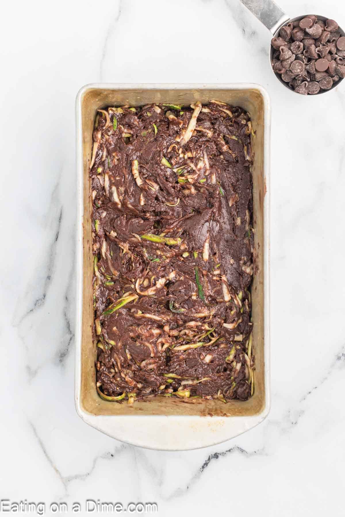 A baking pan brimming with rich chocolate zucchini bread batter rests on a marble surface, while a measuring cup overflowing with chocolate chips sits nearby.