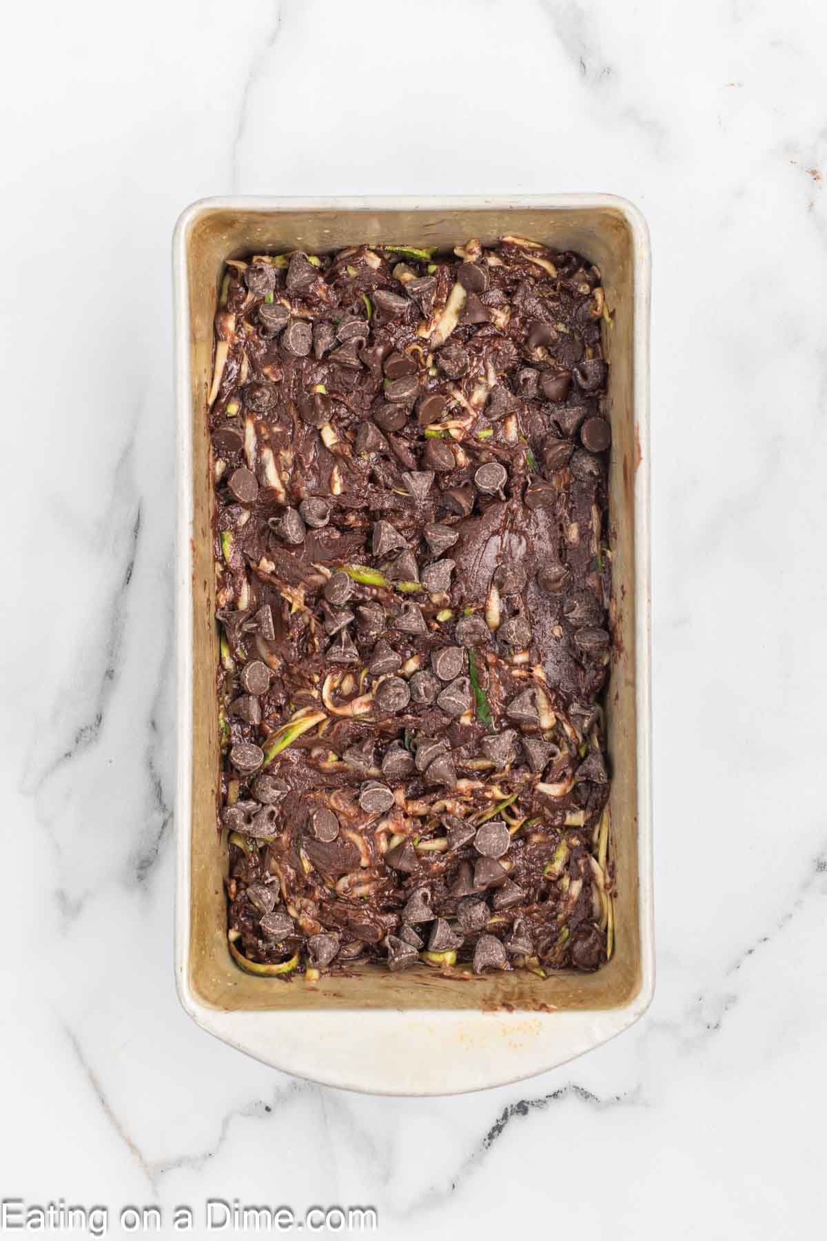 A loaf pan brimming with unbaked chocolate zucchini bread batter, generously topped with chocolate chips, rests on a marble surface. The rich, dark chocolate batter is beautifully speckled with shreds of green zucchini.