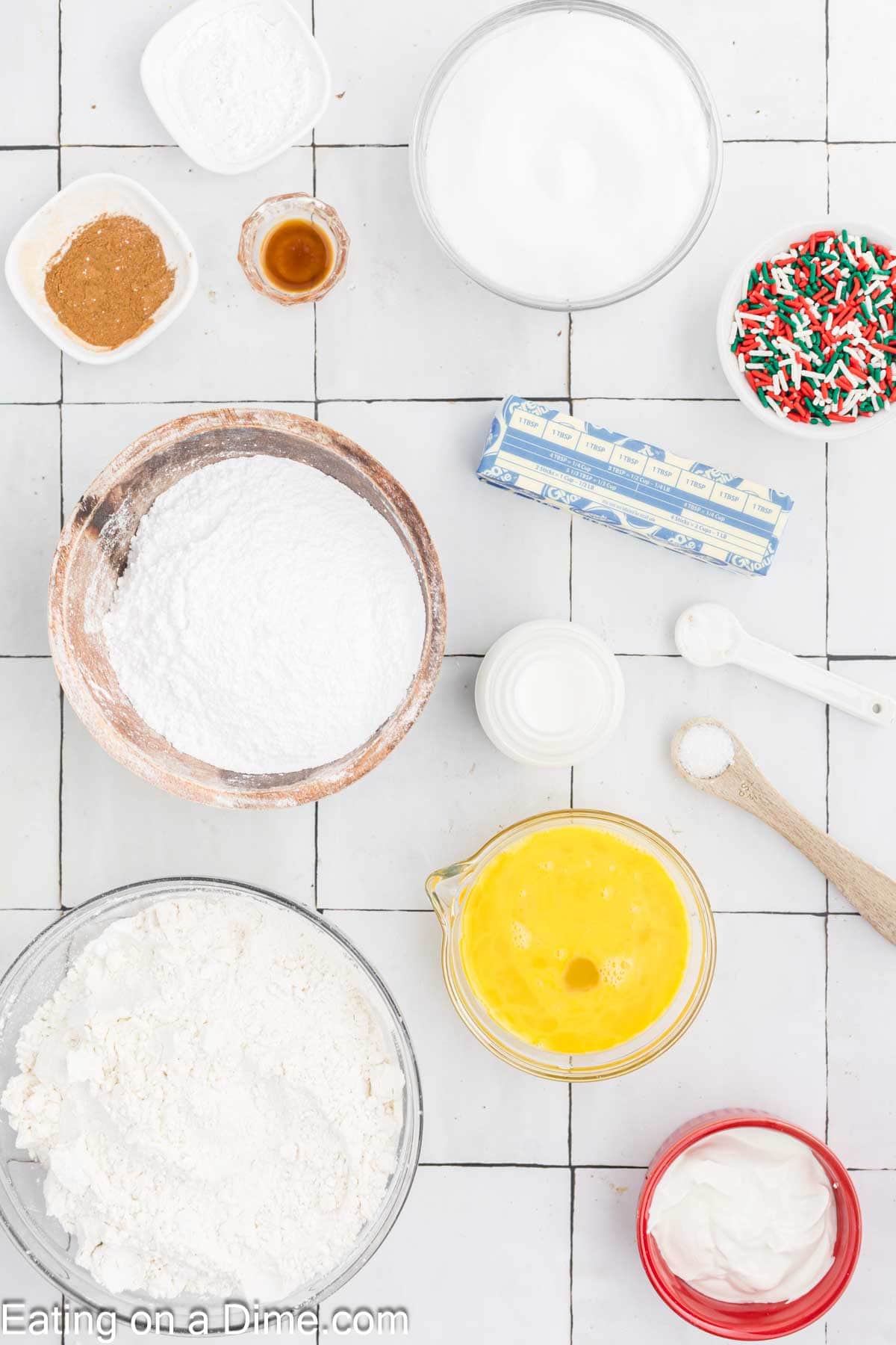 Overhead view of baking ingredients on a tiled surface: flour, powdered sugar, beaten eggs, butter stick, milk, and vanilla extract. A bowl of festive sprinkles hints at a Christmas touch amidst small dishes with baking soda and salt—a perfect setup for holiday bread creation.