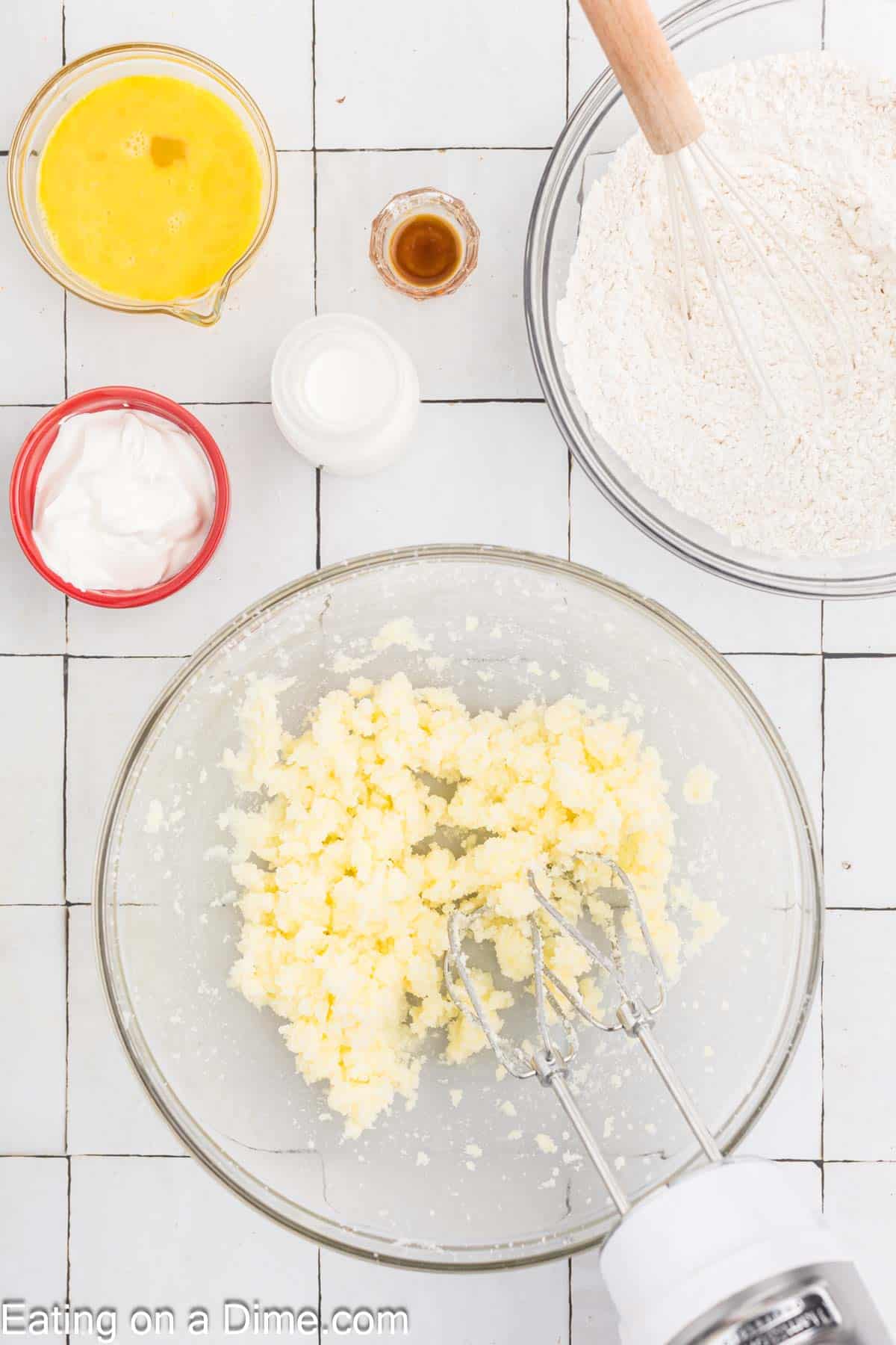 Ingredients for holiday baking are arranged on a tiled surface. There are beaten eggs, flour with a whisk, vanilla extract, milk, and sour cream. A large mixing bowl with creamed butter and sugar is mixed with a hand mixer—perfect for crafting festive bread this Christmas season.