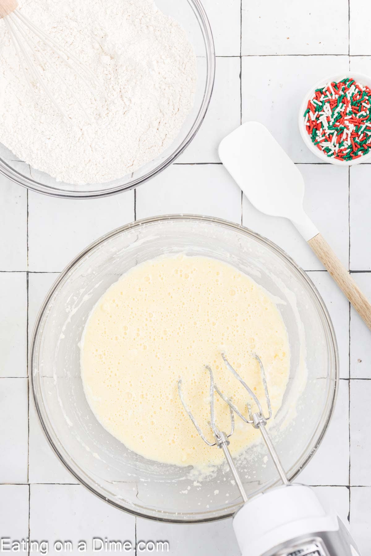 A mixing bowl with beaten batter and a hand mixer sits on a tiled surface, perfect for baking Christmas treats. Nearby, a bowl of flour and a bowl of colorful sprinkles add festive flair. A white spatula rests beside the bowls, ready to create something delightful.