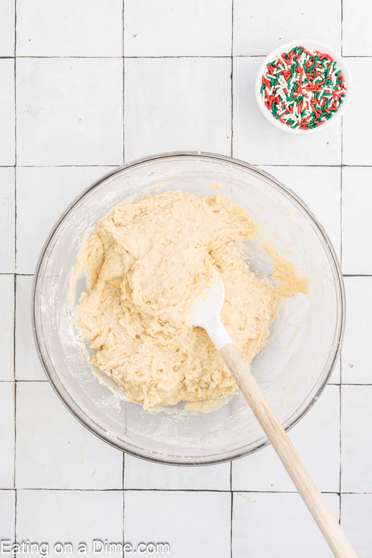 A glass bowl with cookie dough and a spatula rests on a white tiled surface, evoking Christmas warmth. Nearby, a small bowl of colorful sprinkles waits to add holiday cheer.