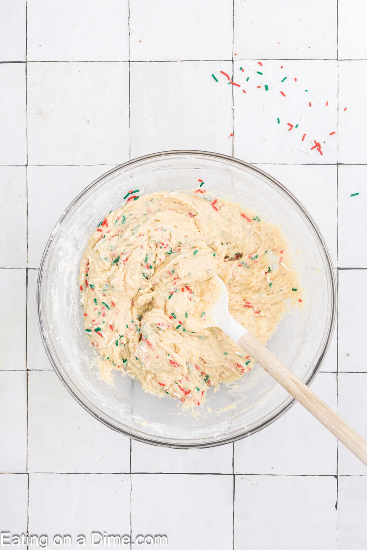 A glass bowl filled with cookie dough mixed with colorful sprinkles sits on a white tiled surface, exuding a cheerful Christmas vibe. A wooden spoon is inside the bowl, and a few festive sprinkles are scattered like holiday joy on the tiles.