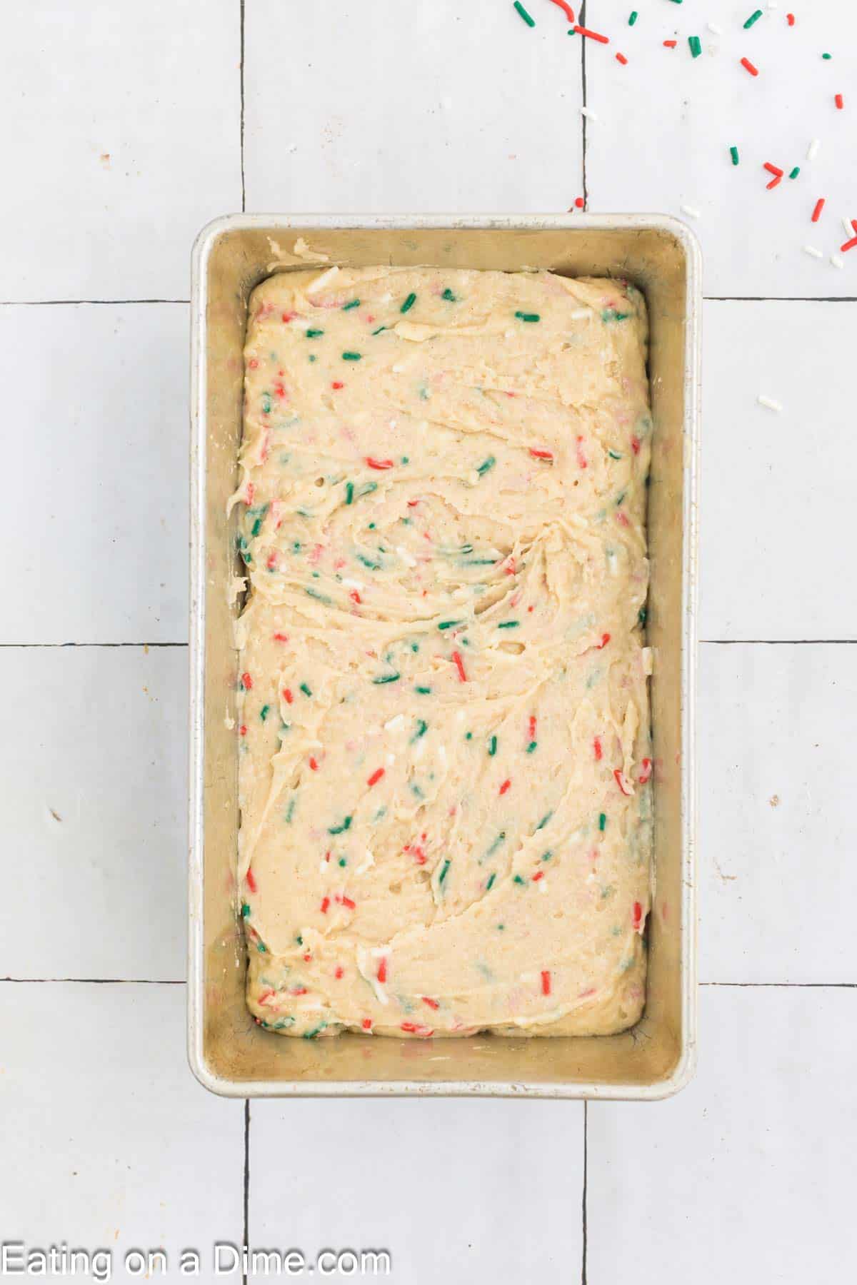 A rectangular baking pan filled with unbaked cake batter mixed with festive red and green sprinkles sits like a Christmas treat on a white tiled surface, surrounded by a few stray sprinkles.