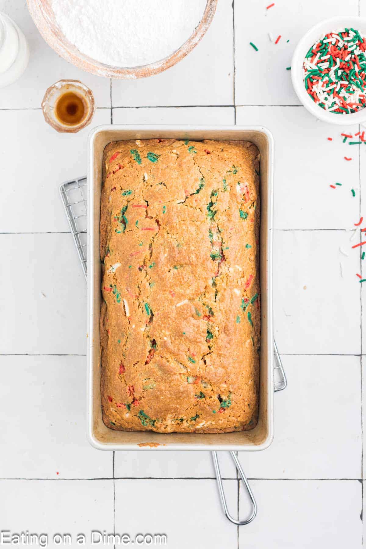 A festive confetti bread loaf sits in a rectangular pan on a cooling rack, evoking Christmas cheer. Nearby, a bowl of powdered sugar, vanilla extract, and vibrant sprinkles adorn the white tiled surface.