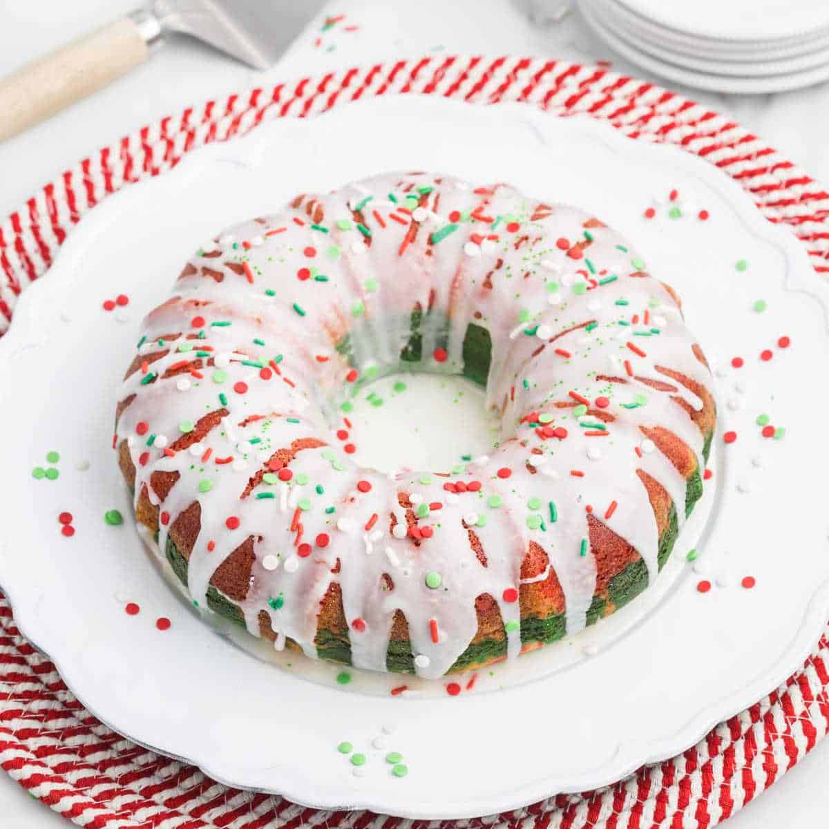 A festive Christmas bundt cake with green and red swirls is topped with white icing and vibrant sprinkles. It rests on a white plate over a red and white woven mat, accompanied by a stack of plates and a cake server in the background, ready to spread holiday cheer.