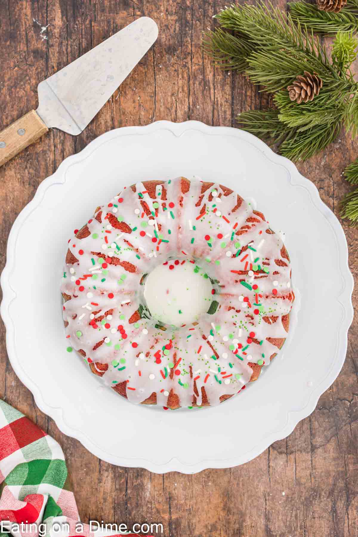 A Christmas bundt cake with white icing and red and green sprinkles sits on a white plate, capturing the holiday spirit. A cake server waits beside it on a wooden table, surrounded by pine branches and a checkered cloth.