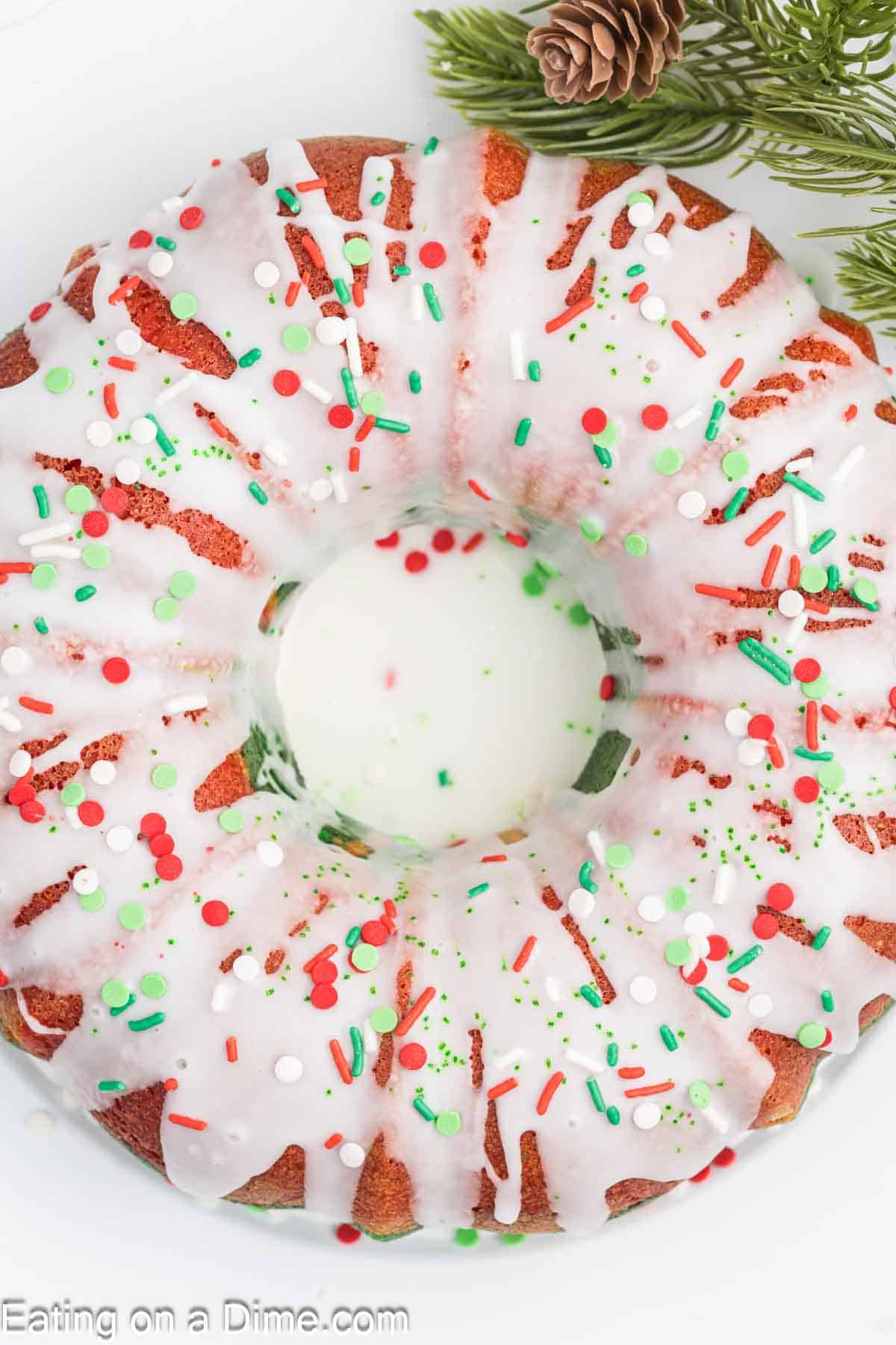 A Christmas bundt cake decorated with white icing and vibrant red, green, and white sprinkles. Pine branches and a small pinecone in the background enhance the joyful holiday theme.