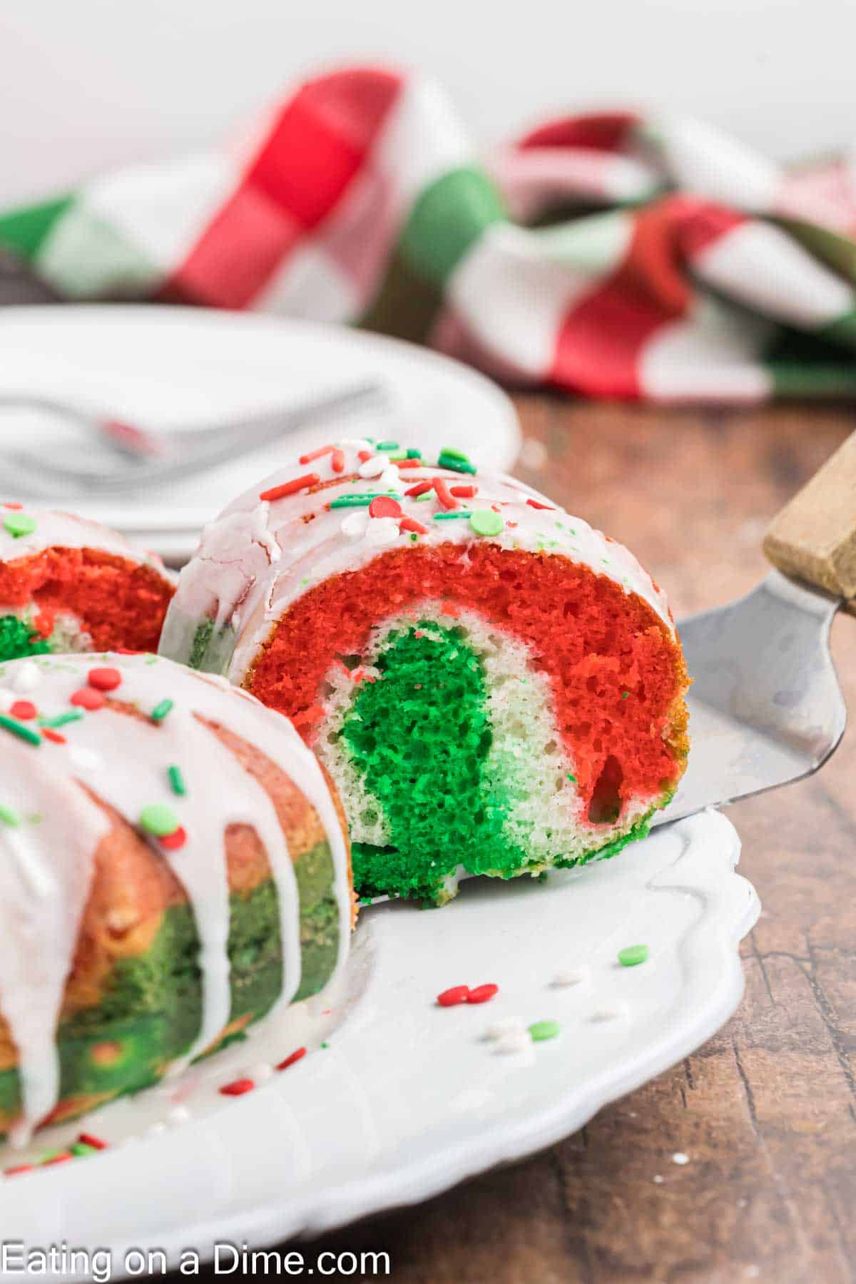 A festive Christmas bundt cake with red, white, and green layers sits on a white plate, topped with a drizzling of white glaze and colorful sprinkles. A knife lifts a slice, ready to serve. In the background, a red, green, and white cloth adds to the holiday cheer.