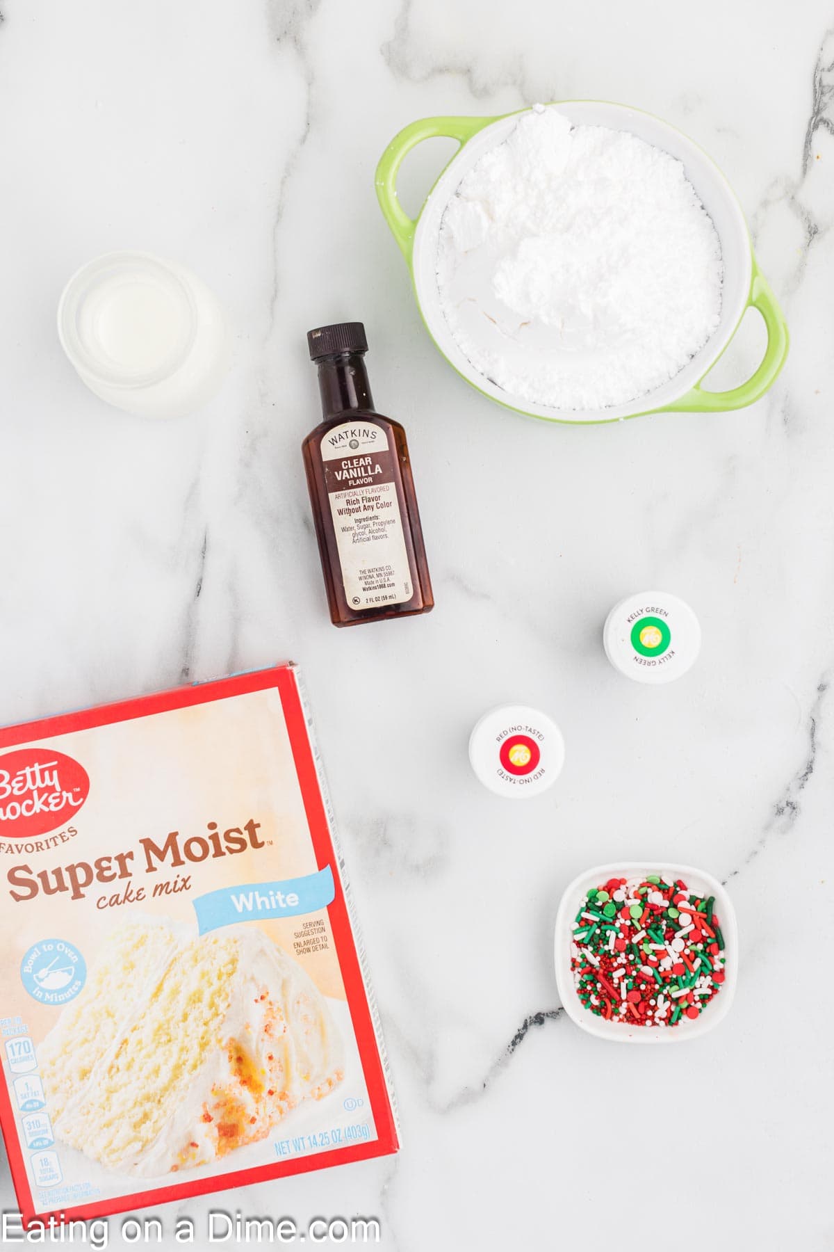 A flat lay of baking items: a box of white cake mix, a small bottle of vanilla extract, a bowl of powdered sugar, two small containers of food coloring (one red, one green), a bowl of festive sprinkles, and a small measuring cup with liquid—perfect for crafting a Christmas Bundt cake.