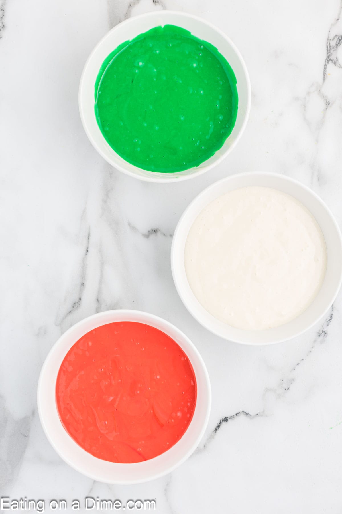 On a marble surface, three white bowls hold festive icing for a Christmas Bundt cake: one brimming with vibrant green, another bursting with merry red, and the last filled with snowy white.