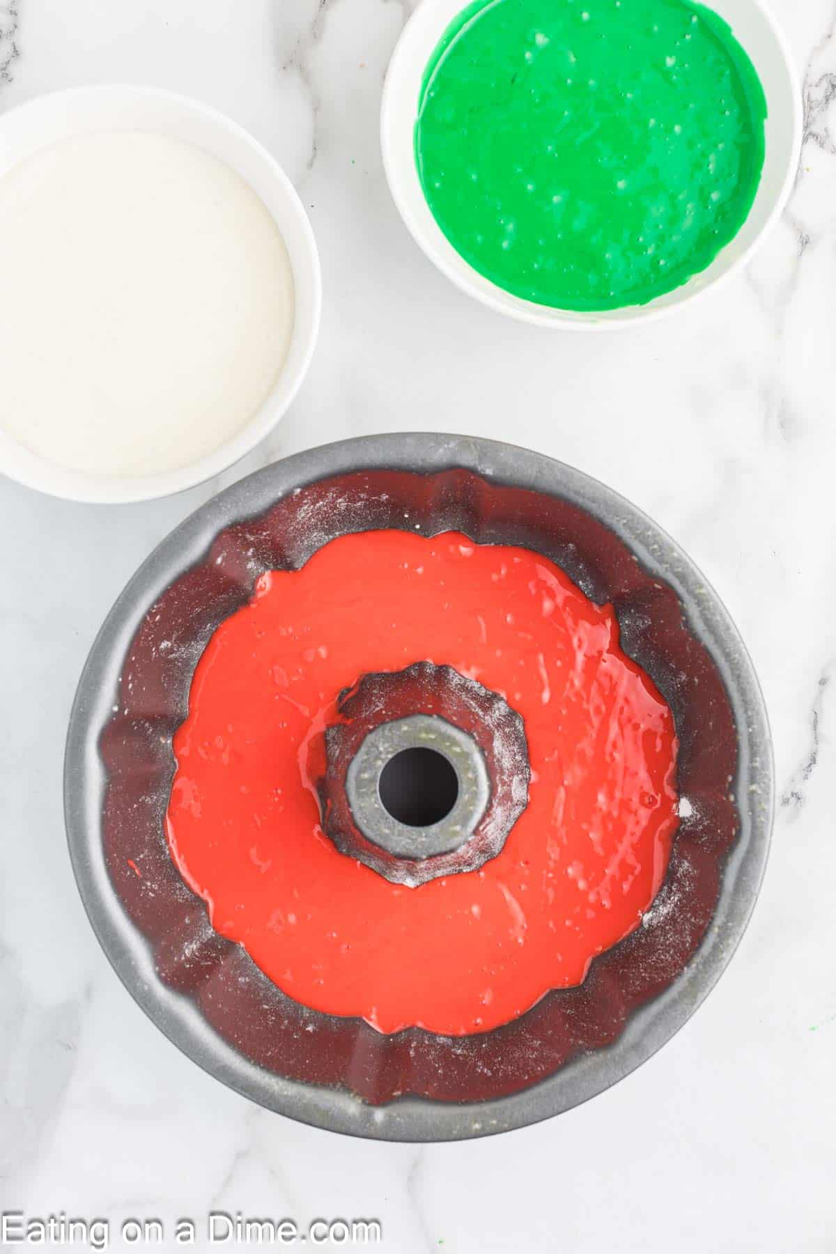 A festive bundt cake pan filled with vibrant red batter sits on a marble countertop, flanked by two bowls brimming with white and green batter, ready to create a delightful Christmas treat.