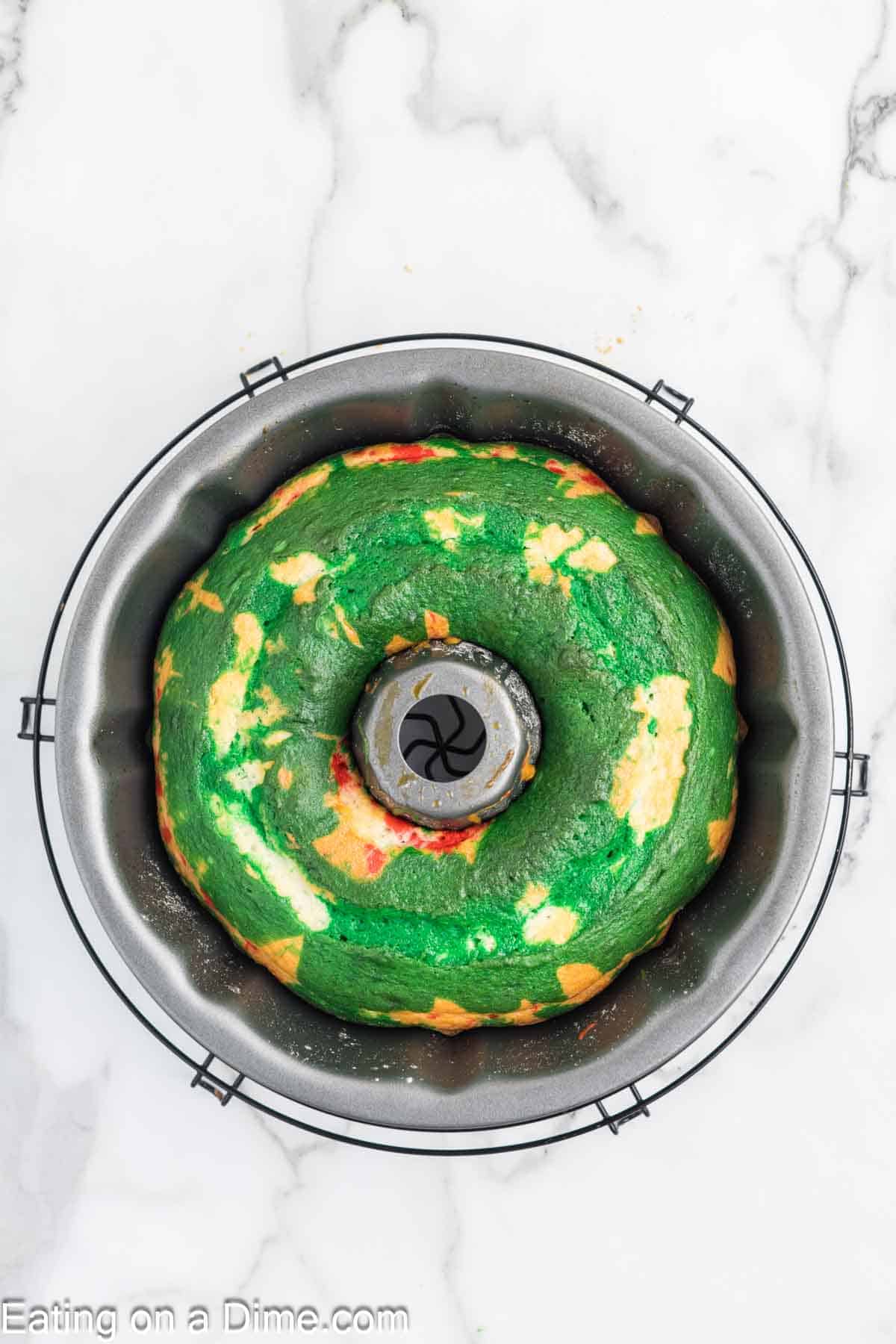 A festive bundt cake with swirled green and white sections sits elegantly inside a round baking pan on a marble surface. Perfect for a Christmas treat, this delightful cake has a charming hole in the center, adding to its seasonal appeal.