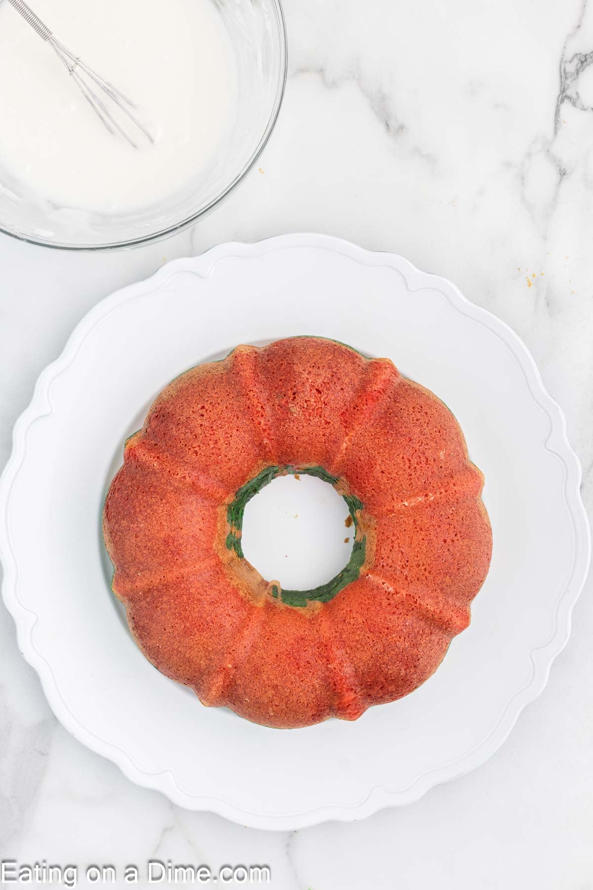 A golden-brown bundt cake sits gracefully on a white plate, its festive allure reminiscent of Christmas. Beside it, a glass bowl brims with rich white icing, ready to add sweetness. The scene rests atop a classic marble countertop, capturing the holiday spirit in every detail.