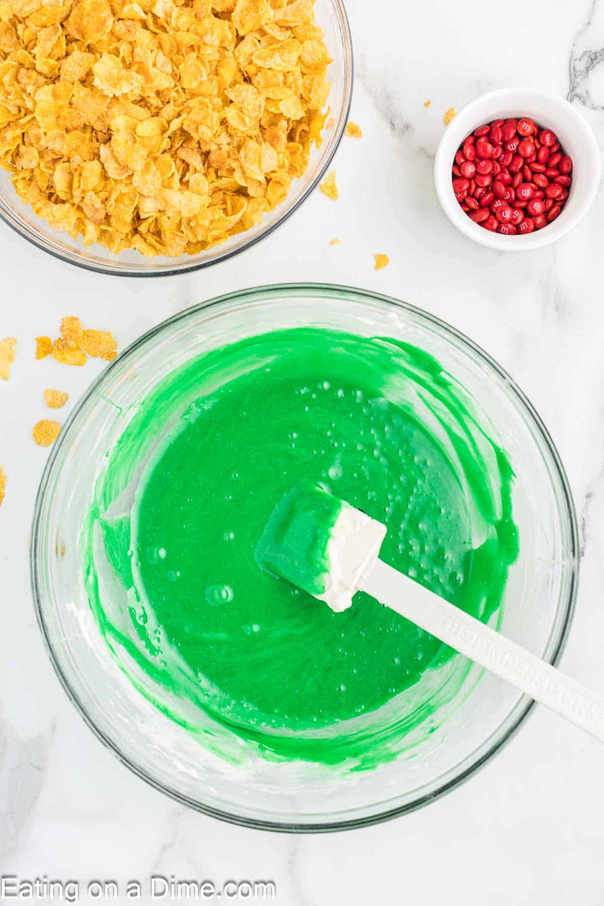 Green melted marshmallow mixture in a bowl with a spatula, with a bowl of corn flakes on the side with a small bowl of red candy on the side