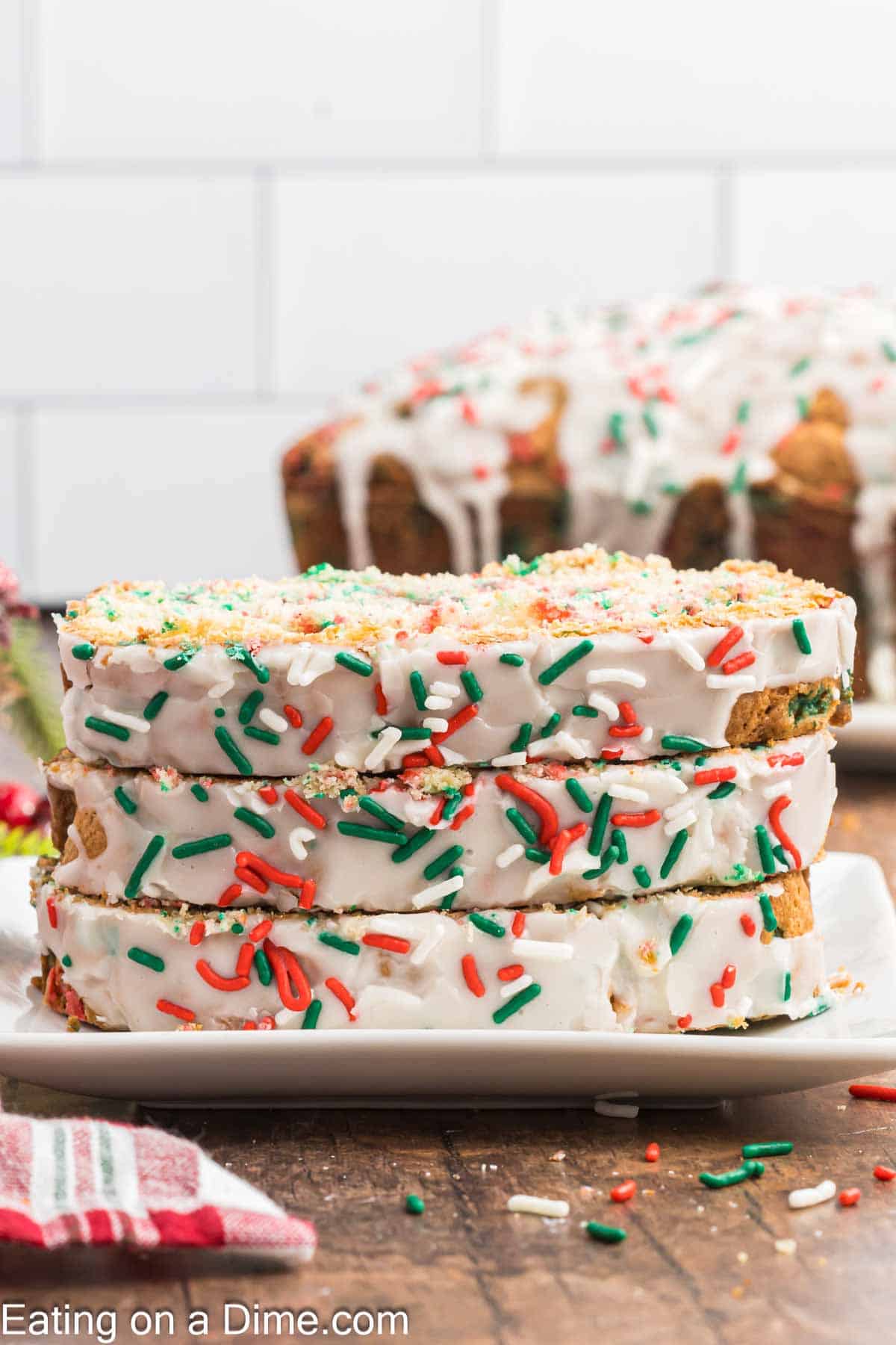 Four slices of festive Christmas bread with white icing and red, green, and white sprinkles are stacked on a plate. Another loaf with similar decoration is blurred in the background, adding to the holiday charm. A kitchen towel is visible to the side.