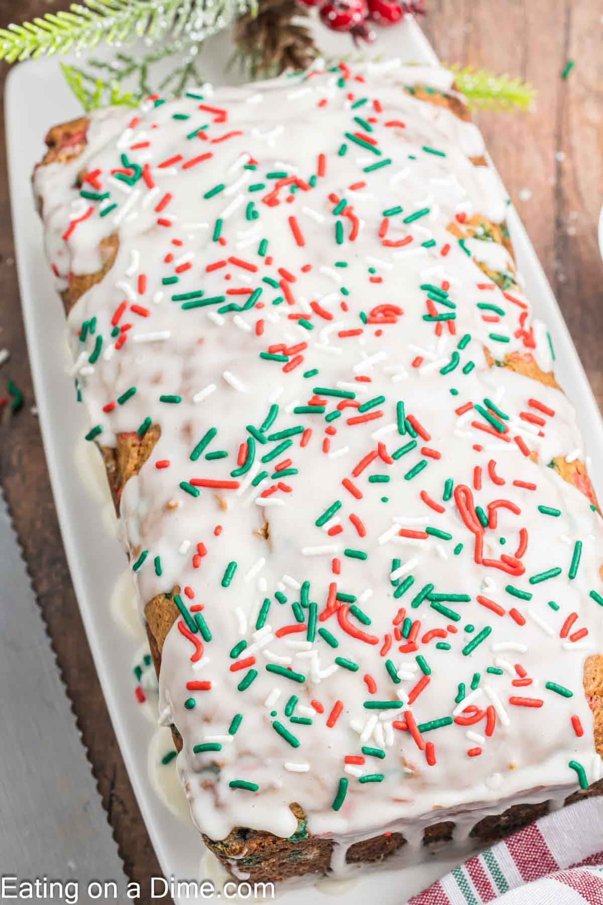 A festive Christmas loaf cake, covered in white icing and adorned with red, green, and white sprinkles, sits invitingly on a rectangular white plate. Holiday-themed decorations, including pine branches, enhance the holiday spirit in the background.