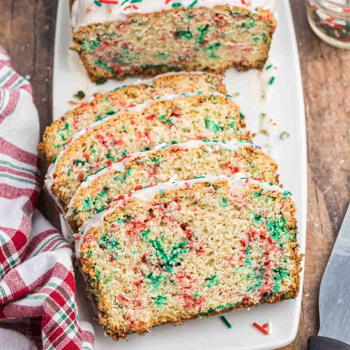 Festive Christmas bread, sliced and adorned with colorful red and green sprinkles, is topped with white icing, elegantly displayed on a white rectangular plate. A red and white checkered napkin rests beside it on the wooden surface.