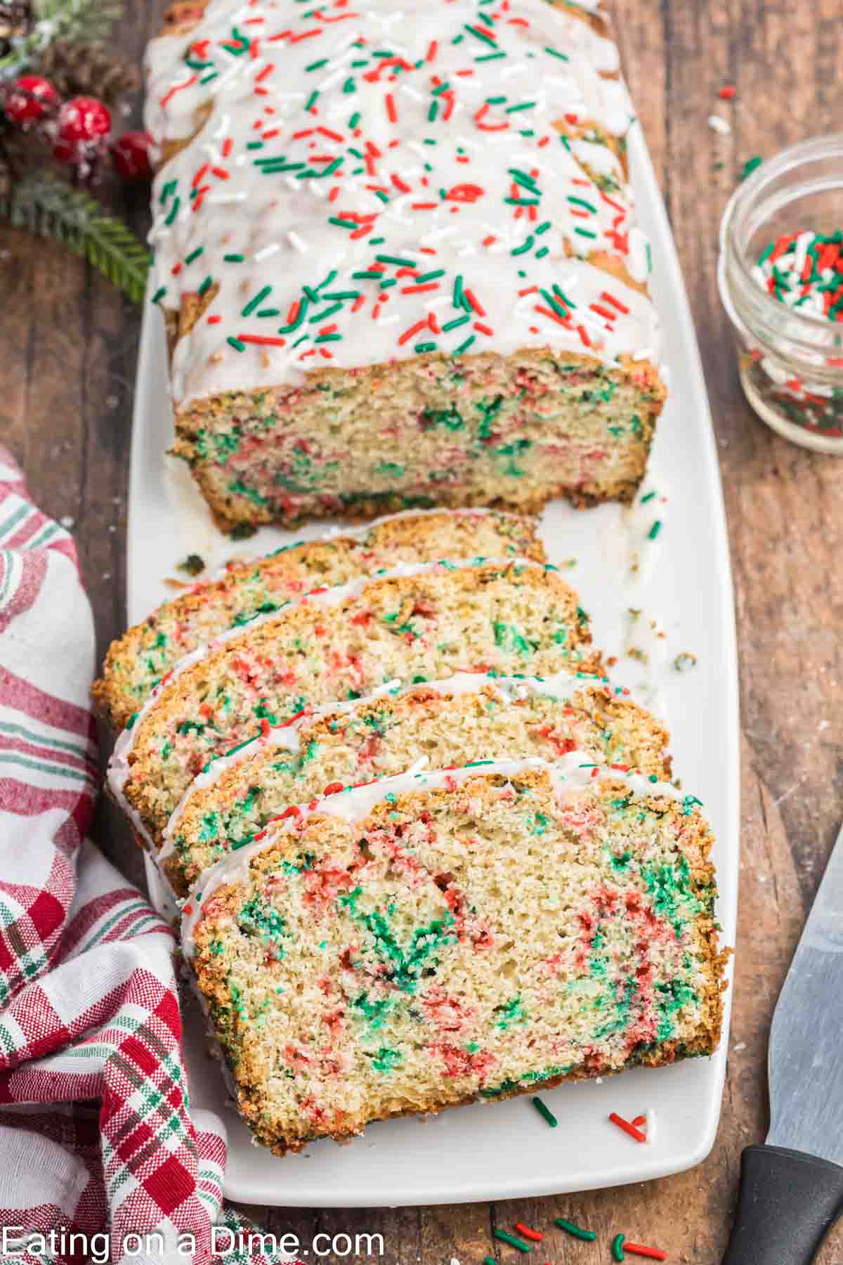 A loaf of festive Christmas bread sits on a white platter, topped with white icing and sprinkled with red and green sprinkles. Several slices are cut, revealing more colorful sprinkles inside. A knife and additional sprinkles are nearby on the wooden surface.