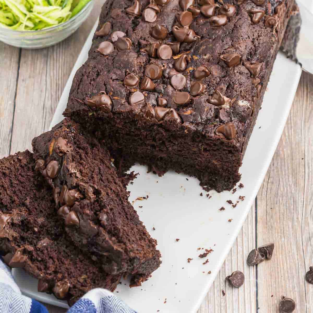 A delightful chocolate zucchini bread topped with chocolate chips rests on a white platter. Several slices have been cut, revealing its moist, rich interior. In the background, a bowl of green zucchini shreds subtly hints at the secret ingredient behind this deliciously indulgent treat.