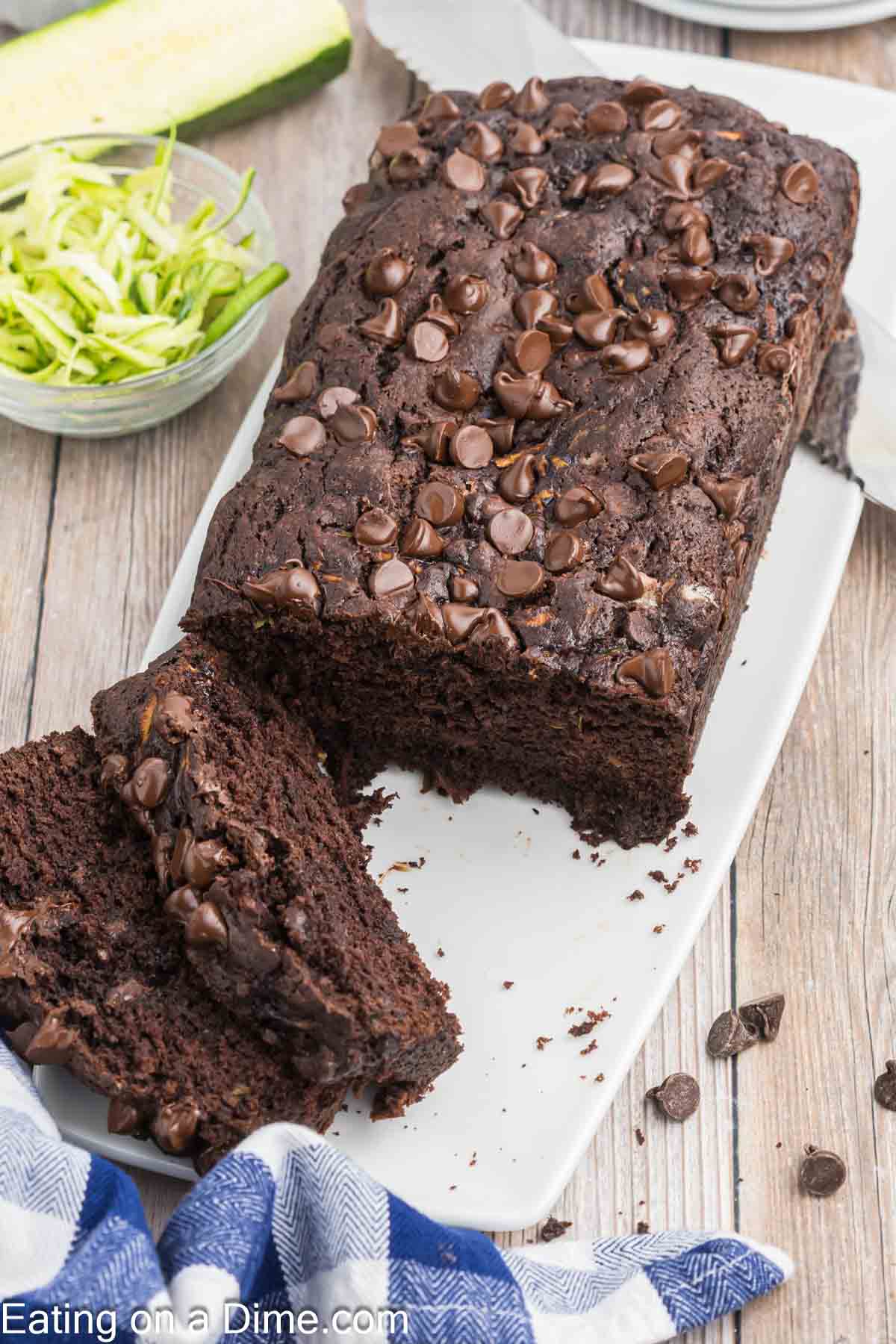 A delicious loaf of chocolate zucchini bread, generously topped with chocolate chips, sits on a white plate. Two slices have been cut from its end, revealing the moist interior. In the background, a bowl of shredded zucchini awaits. A blue and white cloth is partially visible in the corner.