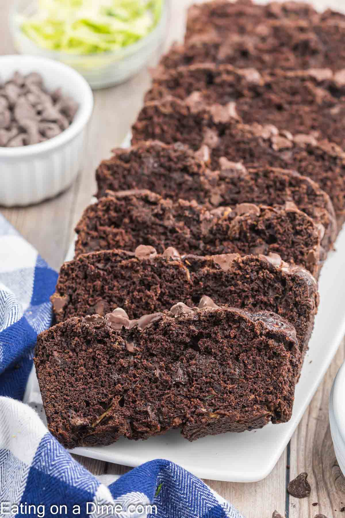 Slices of rich chocolate zucchini bread are neatly arranged on a white rectangular platter. Behind the tempting bread, small bowls brim with chocolate chips and freshly grated zucchini. A blue and white checkered cloth adds a touch of charm to the scene.