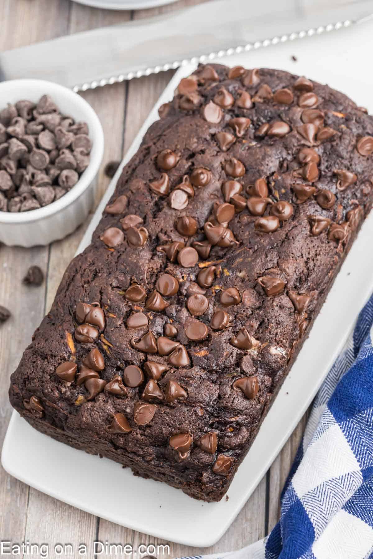 A delicious loaf of chocolate zucchini bread topped with rich chocolate chips rests on a white rectangular plate. Nearby, a bowl brimming with chocolate chips and a striped dish towel complete the inviting scene.