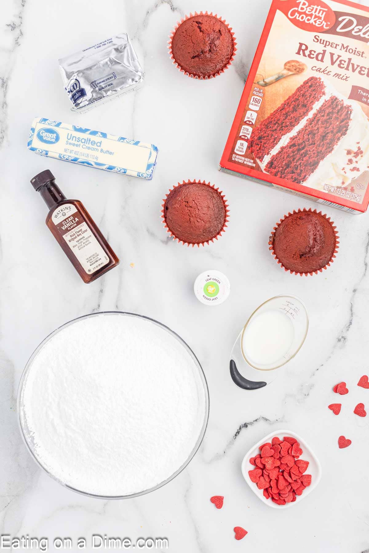 Baking ingredients for festive red velvet cupcakes are laid out on a marble surface. Includes a box of red velvet cake mix, baked cupcakes, sugar, unsalted butter, vanilla extract, milk, and heart-shaped decorations perfect for any celebration—even one as mischievous as the Grinch's.