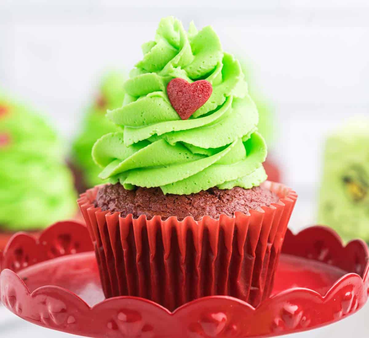 A chocolate cupcake with bright green frosting, whimsically resembling a Grinch-style Christmas tree, is topped with a small red heart. It sits on a decorative red plate, while other similar Grinch-themed cupcakes are blurred in the background.