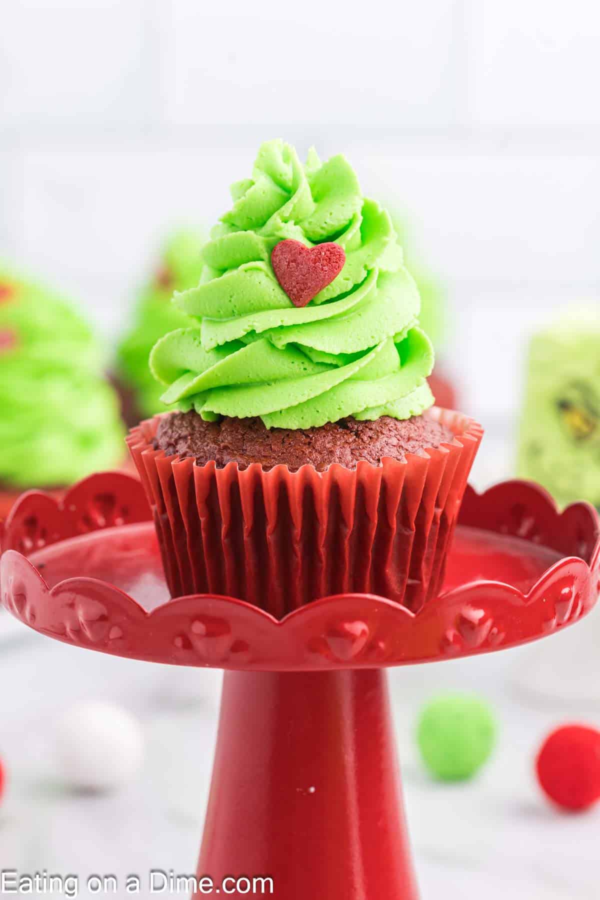 A festive Grinch-themed cupcake with green frosting shaped like a Christmas tree is topped with a red heart decoration, perched on a red cake stand. The cupcake liner matches in red, and blurred decorations create a whimsical background.